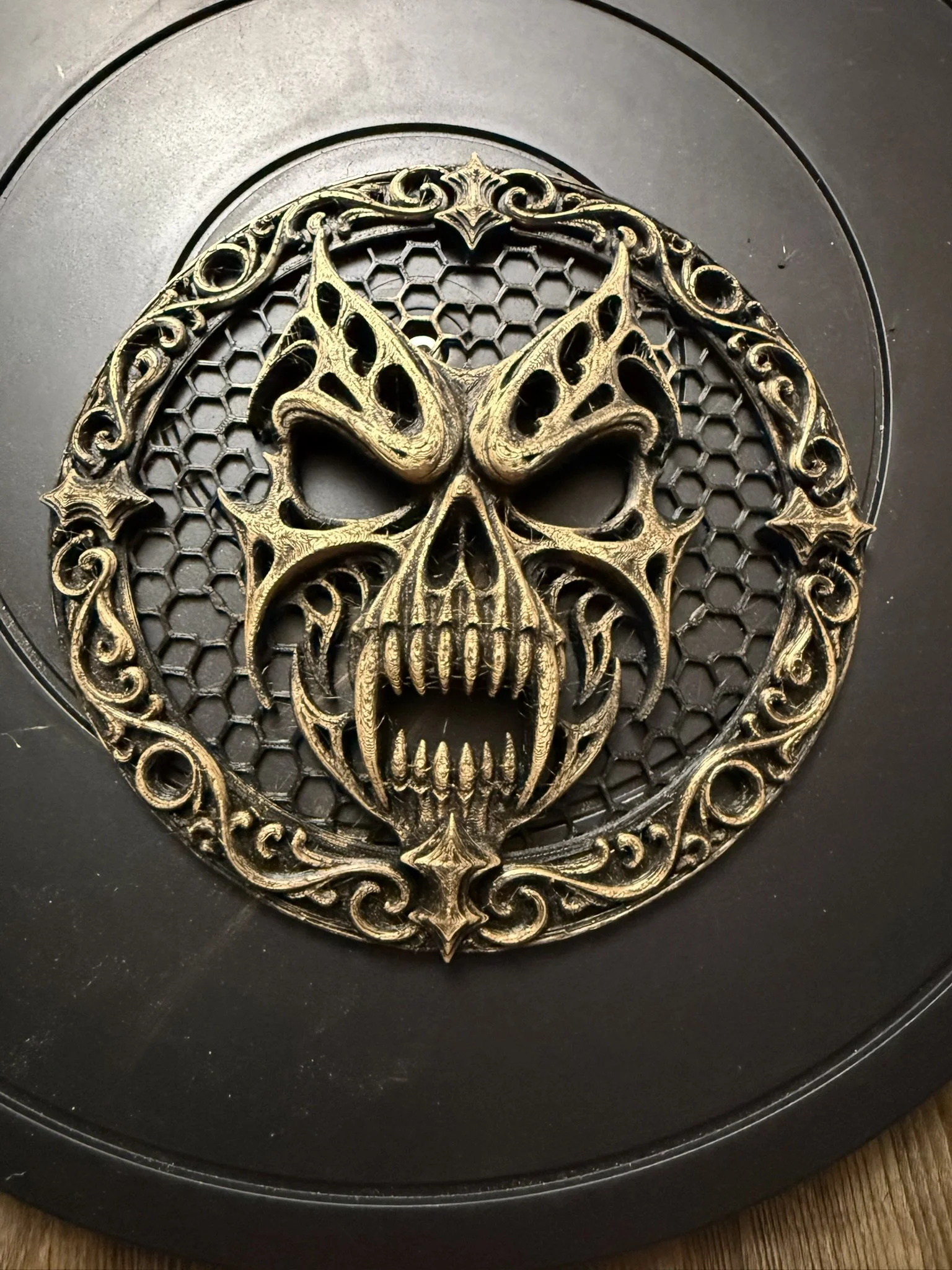 A decorative metal skull with intricate designs on a honeycomb background, surrounded by a circular ornate frame, placed on a black surface.