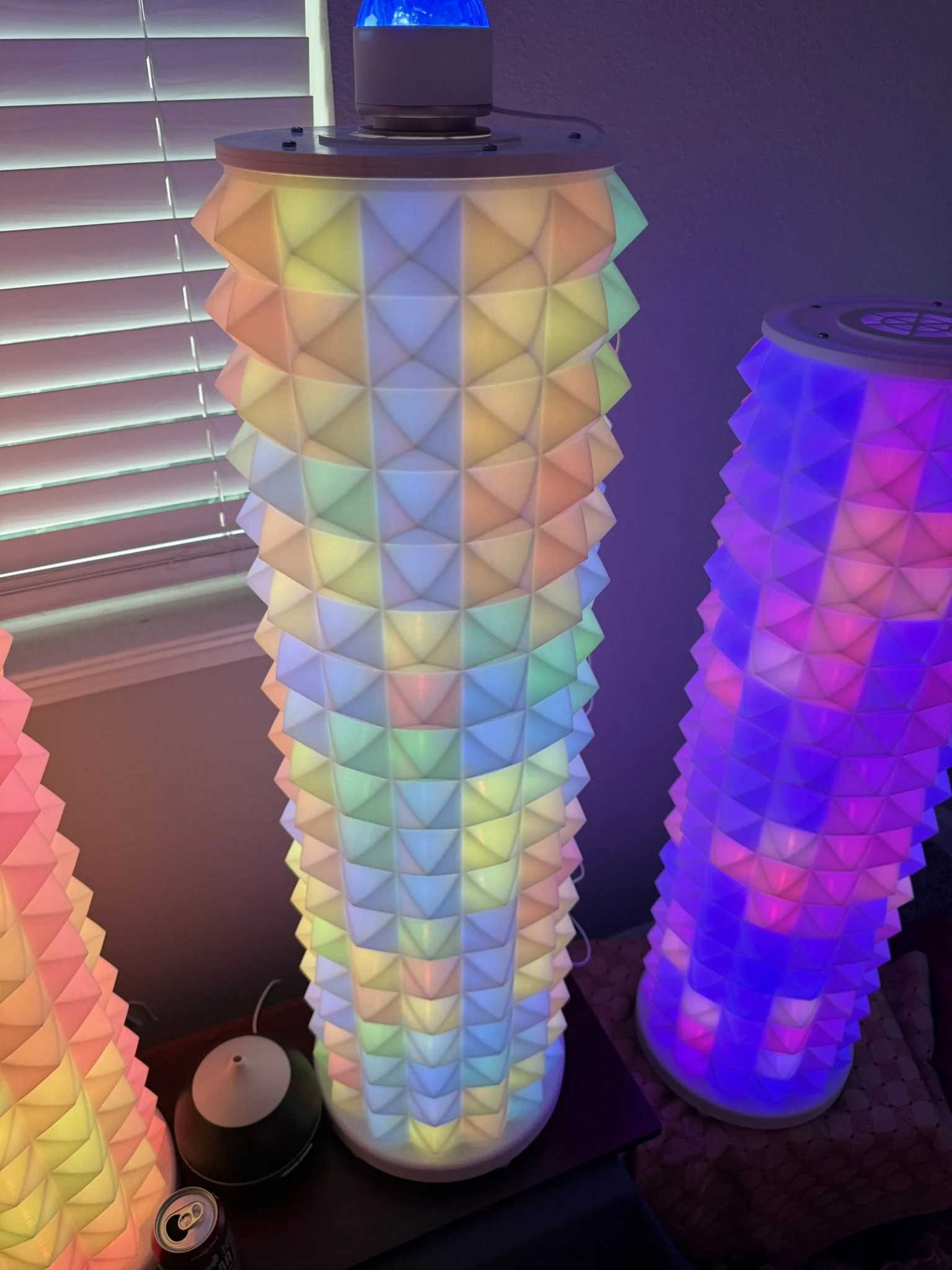 Colorful, geometric, illuminated lamp with layered pointed shapes in pastel colors standing on a table, with another similar blue-lit lamp nearby and a window with blinds in the background.