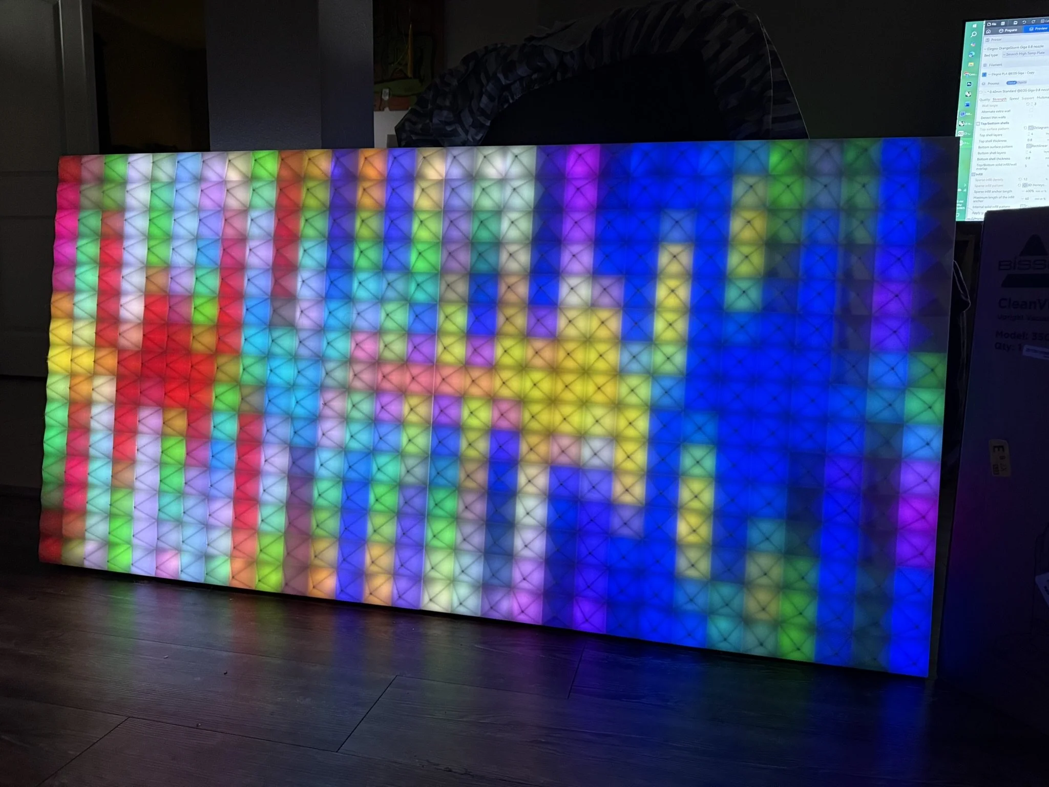 Colorful LED grid with rainbow pattern leaning against a wall, seen in a dimly lit room, with a computer monitor visible in the background.