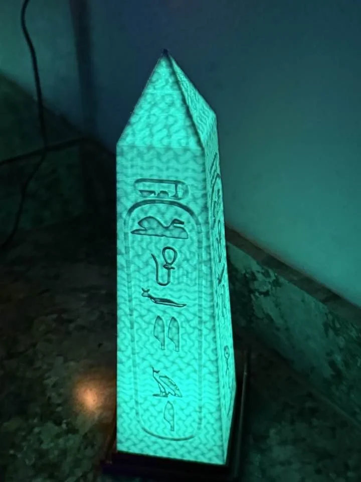 Illuminated Egyptian-themed lamp with hieroglyphs, emitting a teal glow, placed on a dark surface near a wall.