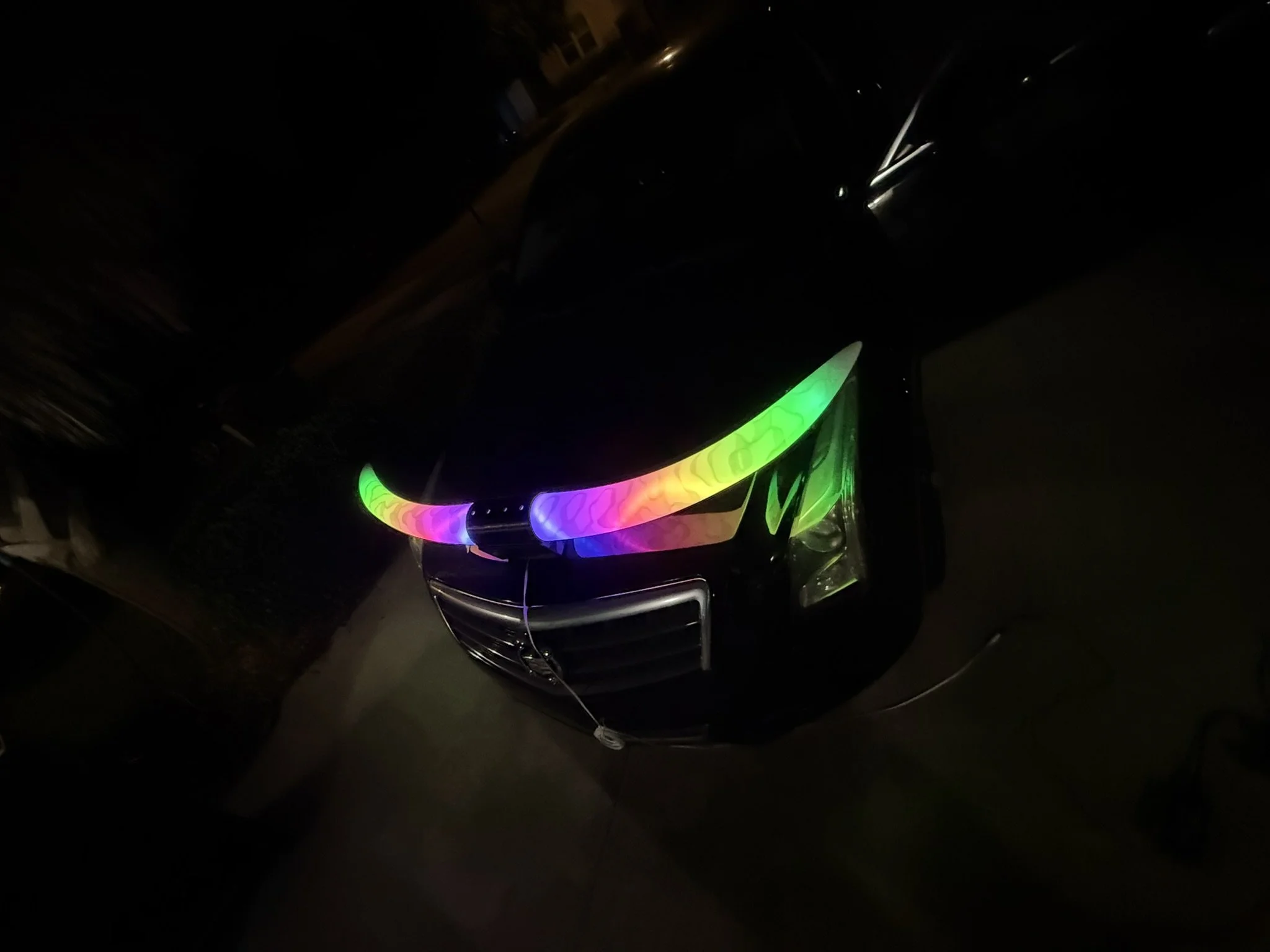 A black car with rainbow-colored LED lights on the front bumper parked in a dark area.