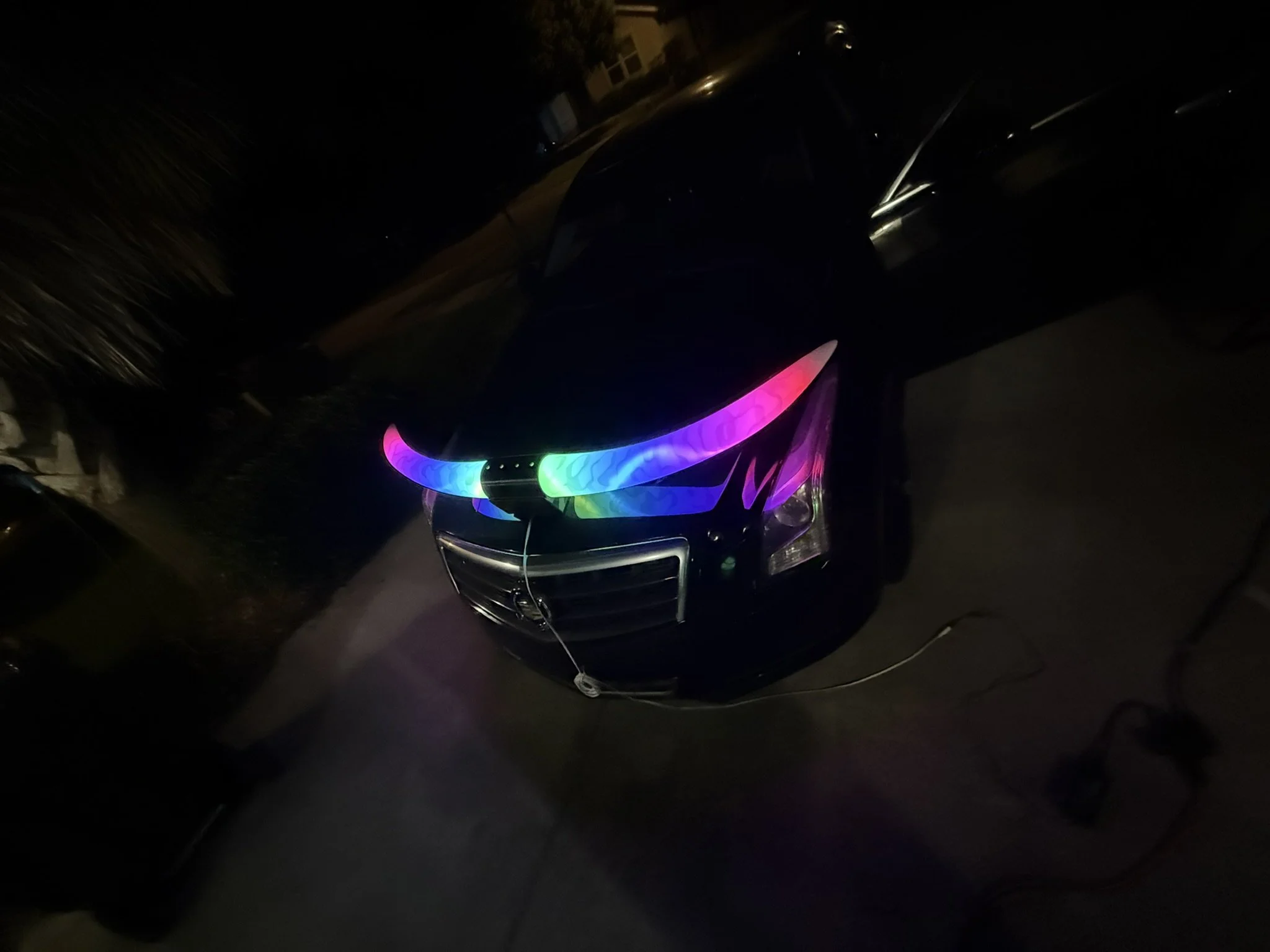 A black car with illuminated, rainbow-colored LED lights on the front grille parked outdoors at night.