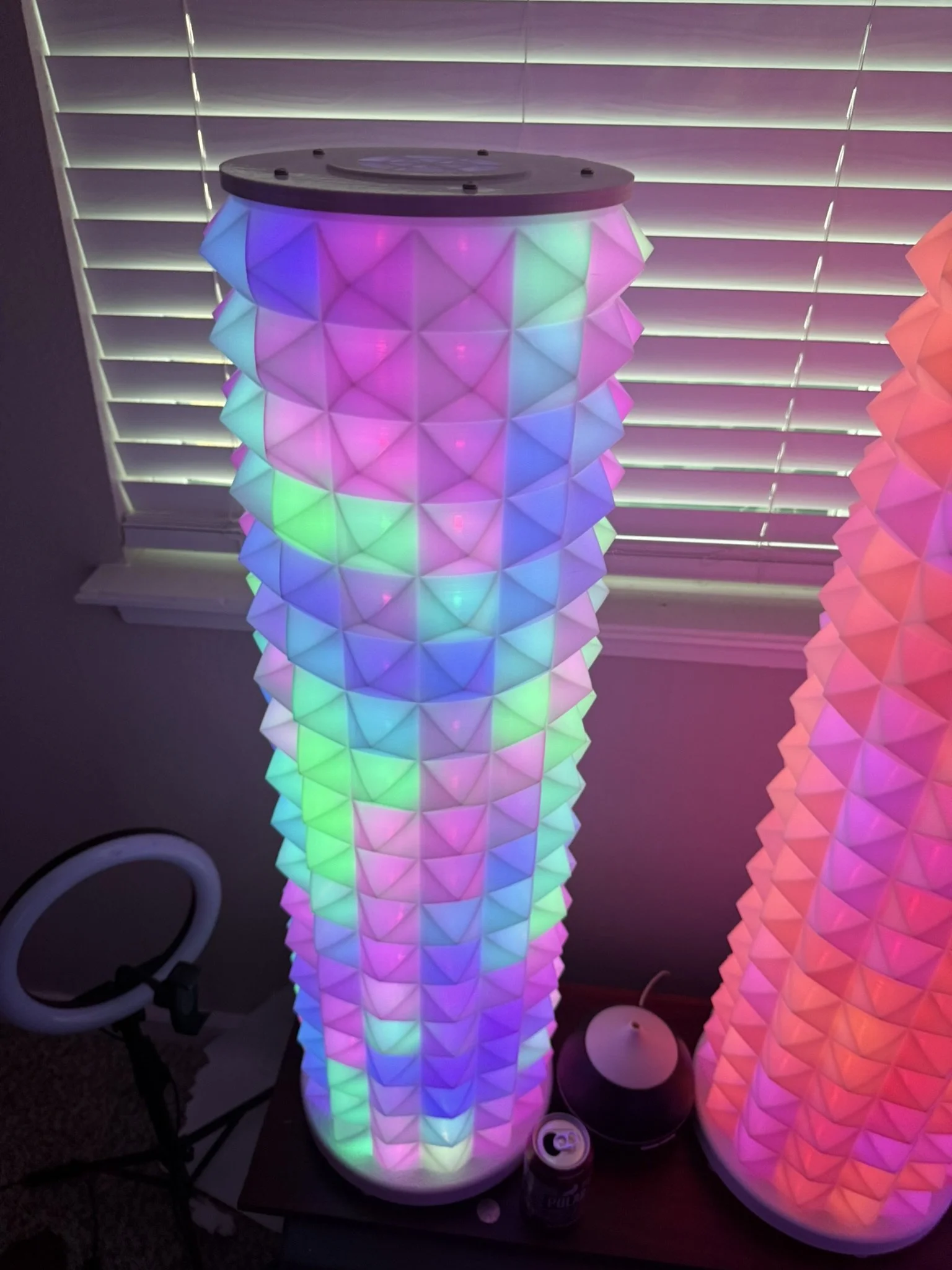 Two tall LED lamps with colorful, geometric, cone-shaped shades glowing in purple, blue, green, and pink hues, placed on a dark surface near a window with closed blinds.