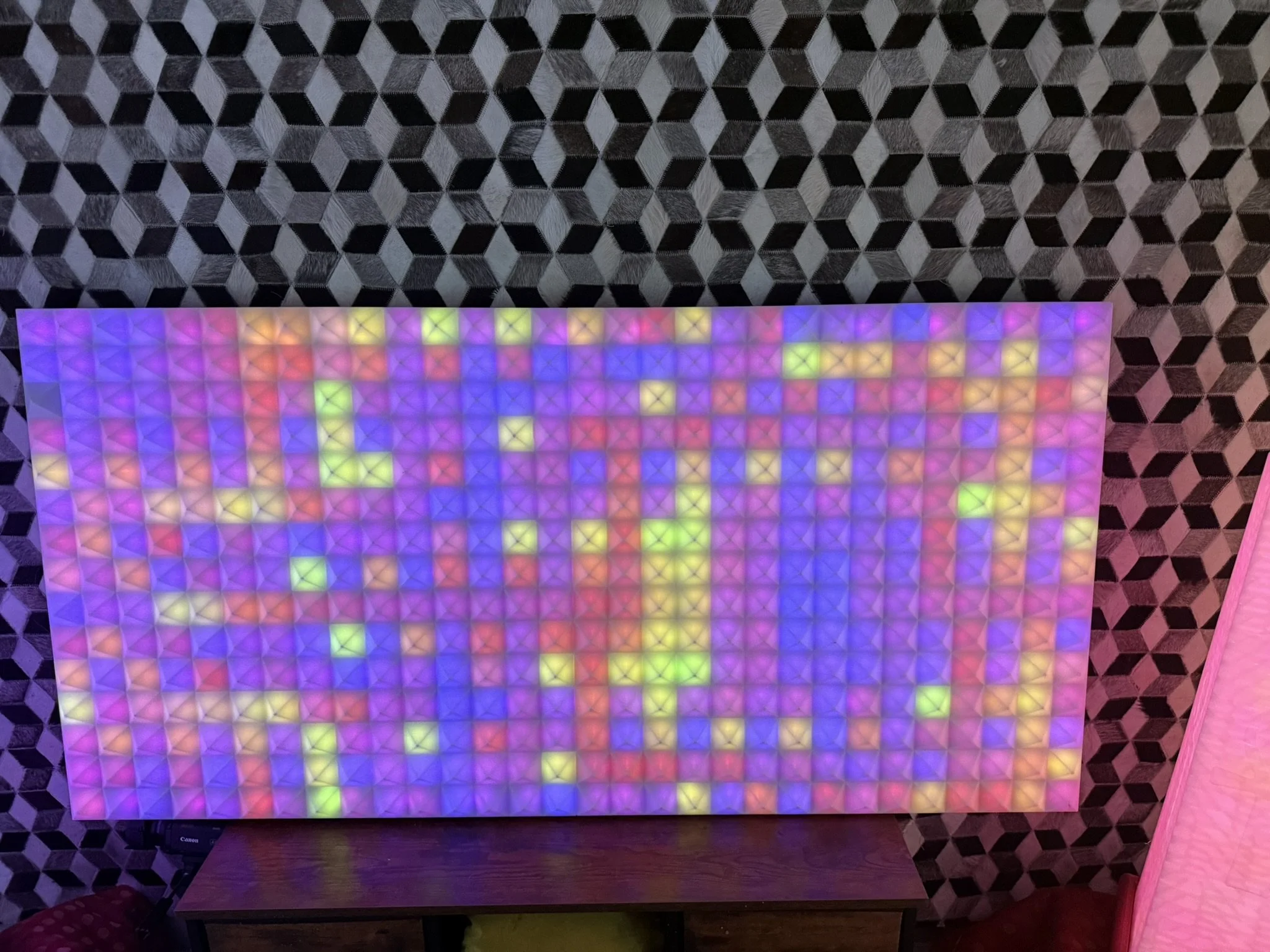 A multicolored LED light panel with a grid of small squares, displaying vibrant colors like pink, yellow, green, blue, and purple, placed on a wooden table against a background of a patterned wall.