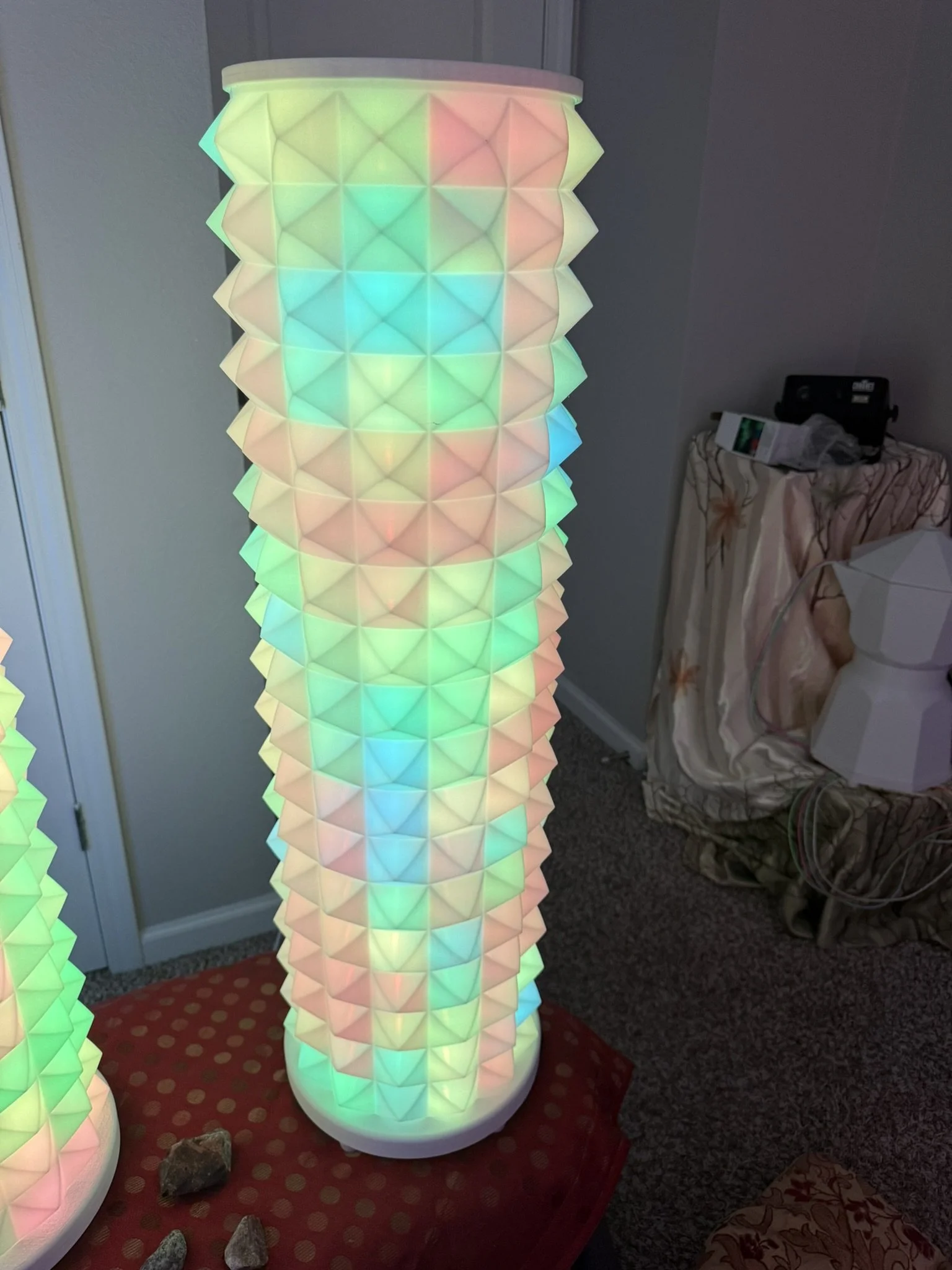 Colorful illuminated lamp with a geometric pattern, situated indoors on a red polka dot mat, with stones nearby and furniture on the side.