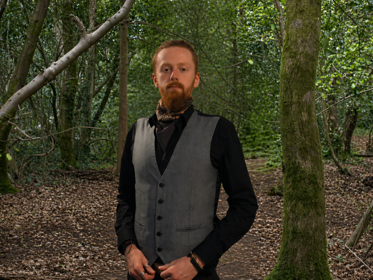 Man with red hair and beard wearing a black shirt, gray vest, and scarf standing in a dense forest.