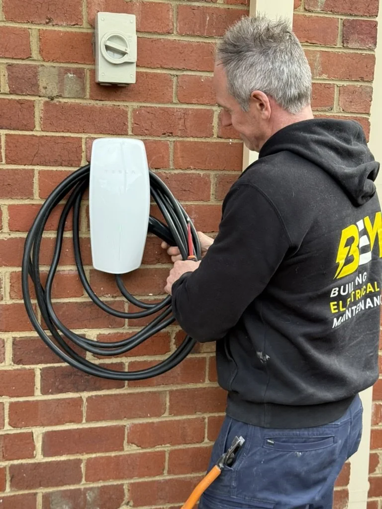 A man with gray hair wearing a black hoodie with yellow text is installing or servicing an electric vehicle charging station on a brick wall.