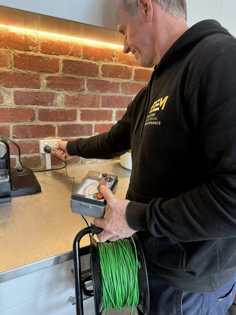 Man in black hoodie holding a testing device and a spool of green wire, testing an electrical outlet in a room with a brick wall.