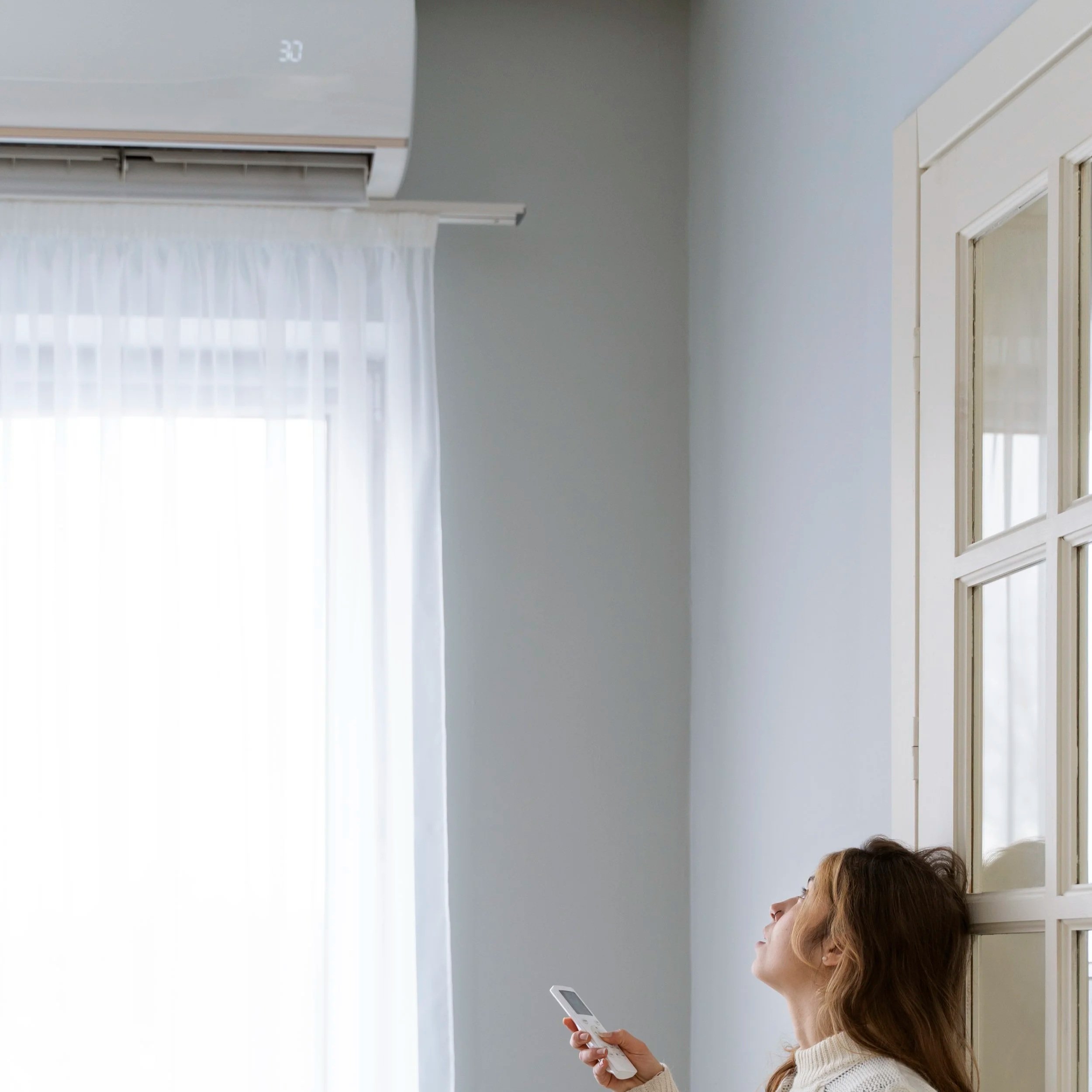A woman with wavy hair holds a remote control and looks up towards an air conditioning unit mounted on the wall above a window with white curtains.