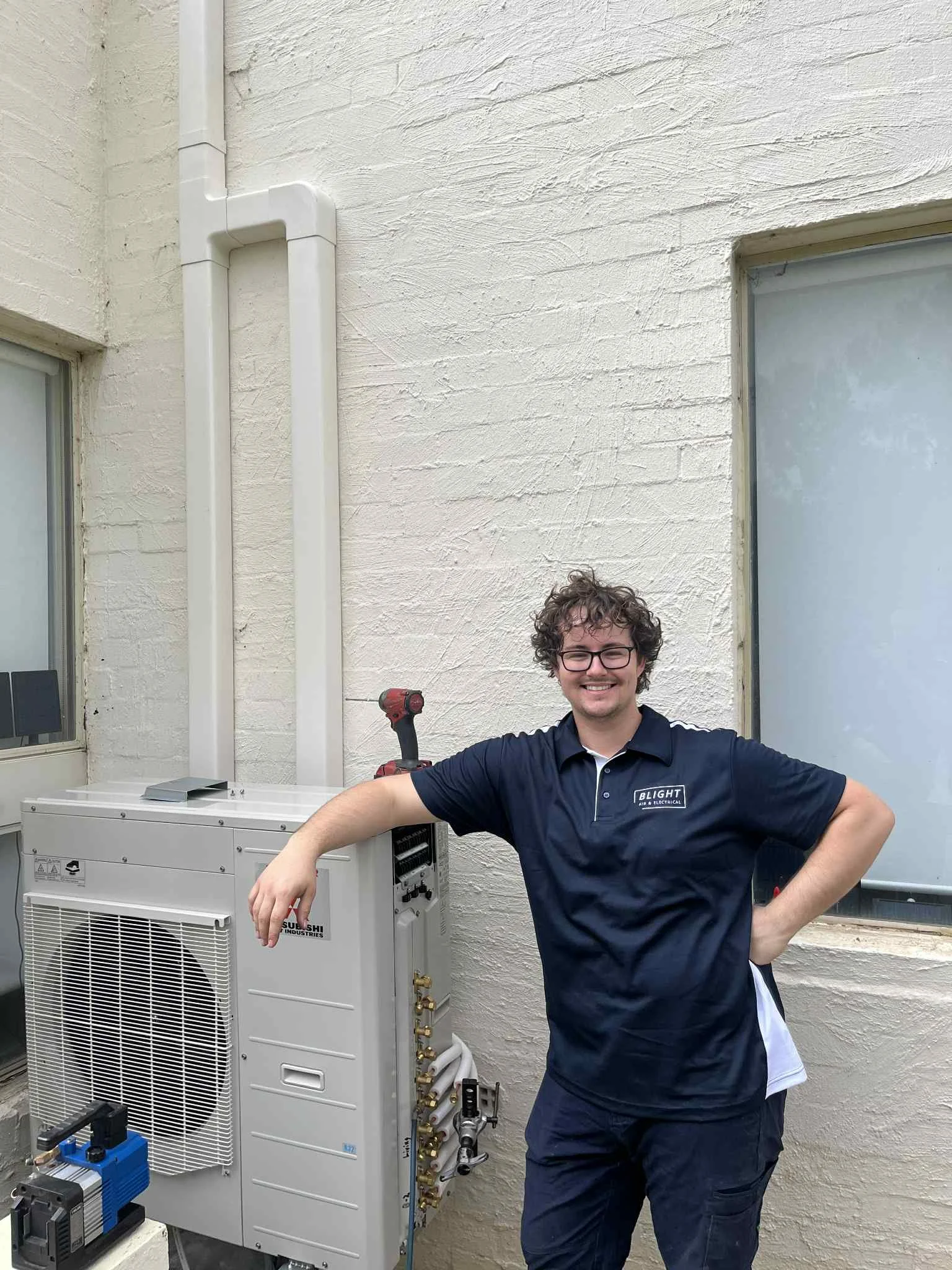 A smiling man with curly hair and glasses, wearing a navy blue polo shirt and navy pants, standing next to a white HVAC or electrical unit outside a building with cream-colored brick walls, a window, and visible pipes.