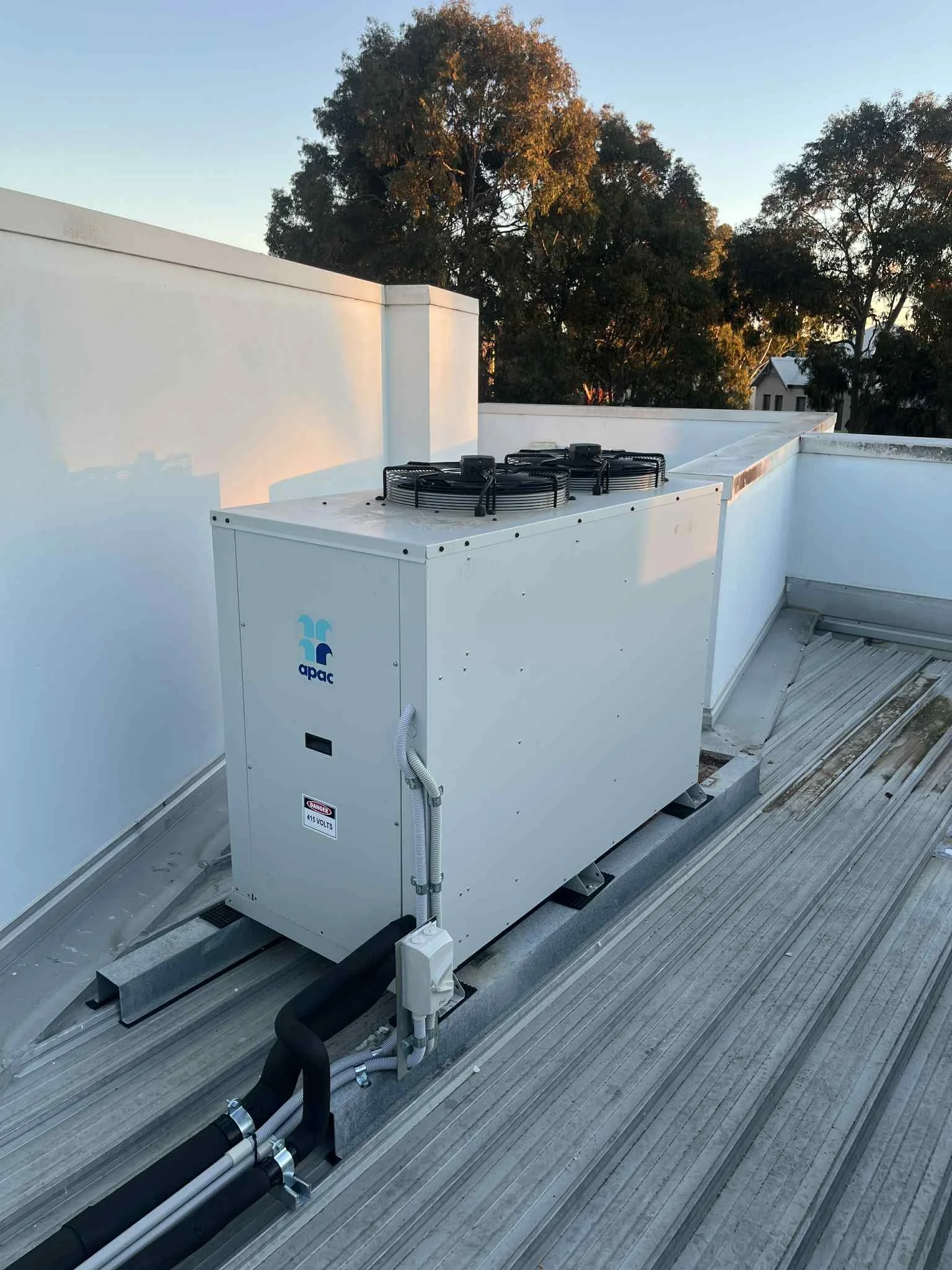 Rooftop HVAC unit with a white exterior, black fans on top, and electrical conduits and pipes connected at the base. The background shows trees and part of a building under a clear sky.