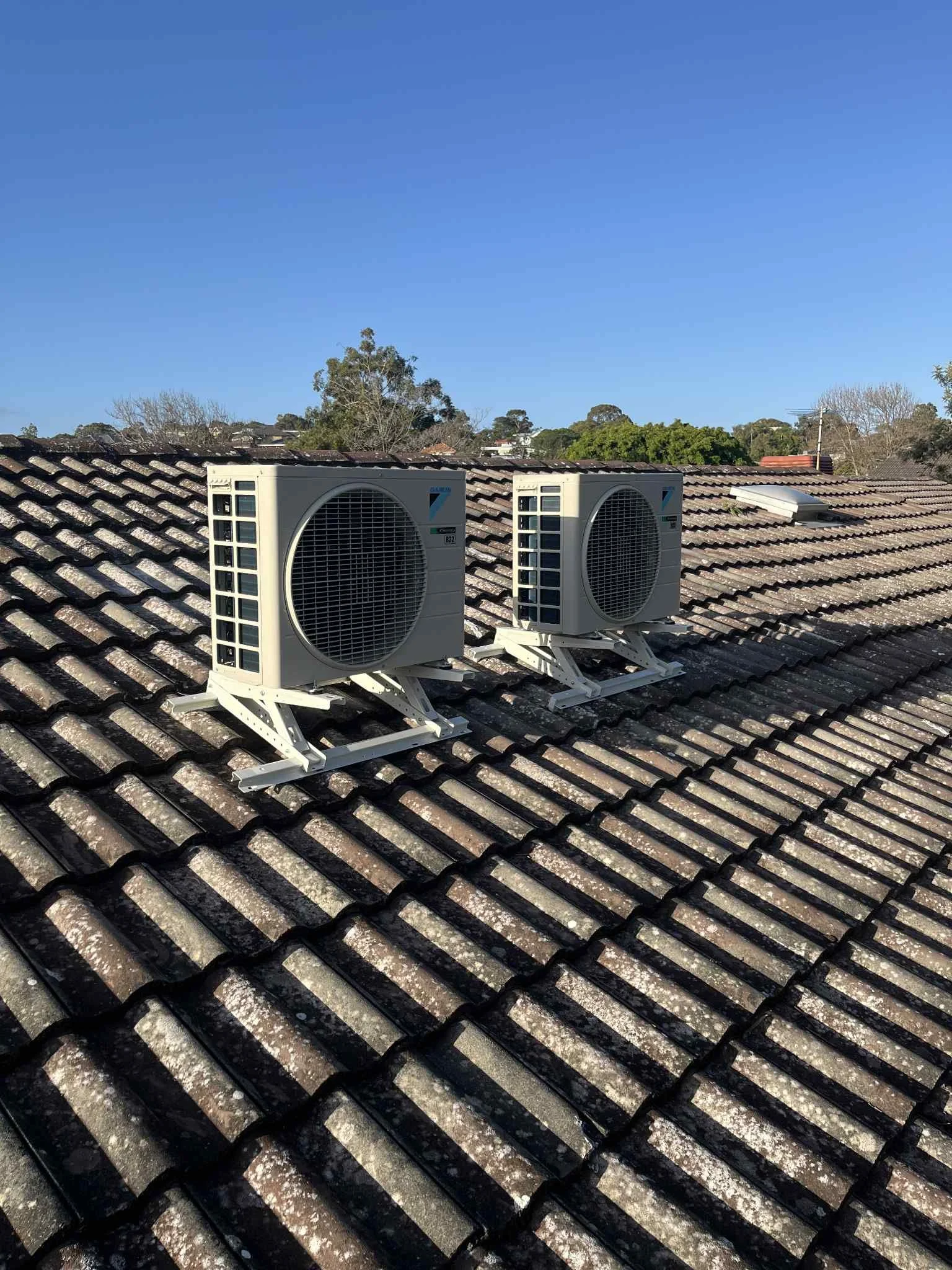 Two outdoor air conditioning units installed on a sloped roof with weathered tiles, under a clear blue sky.