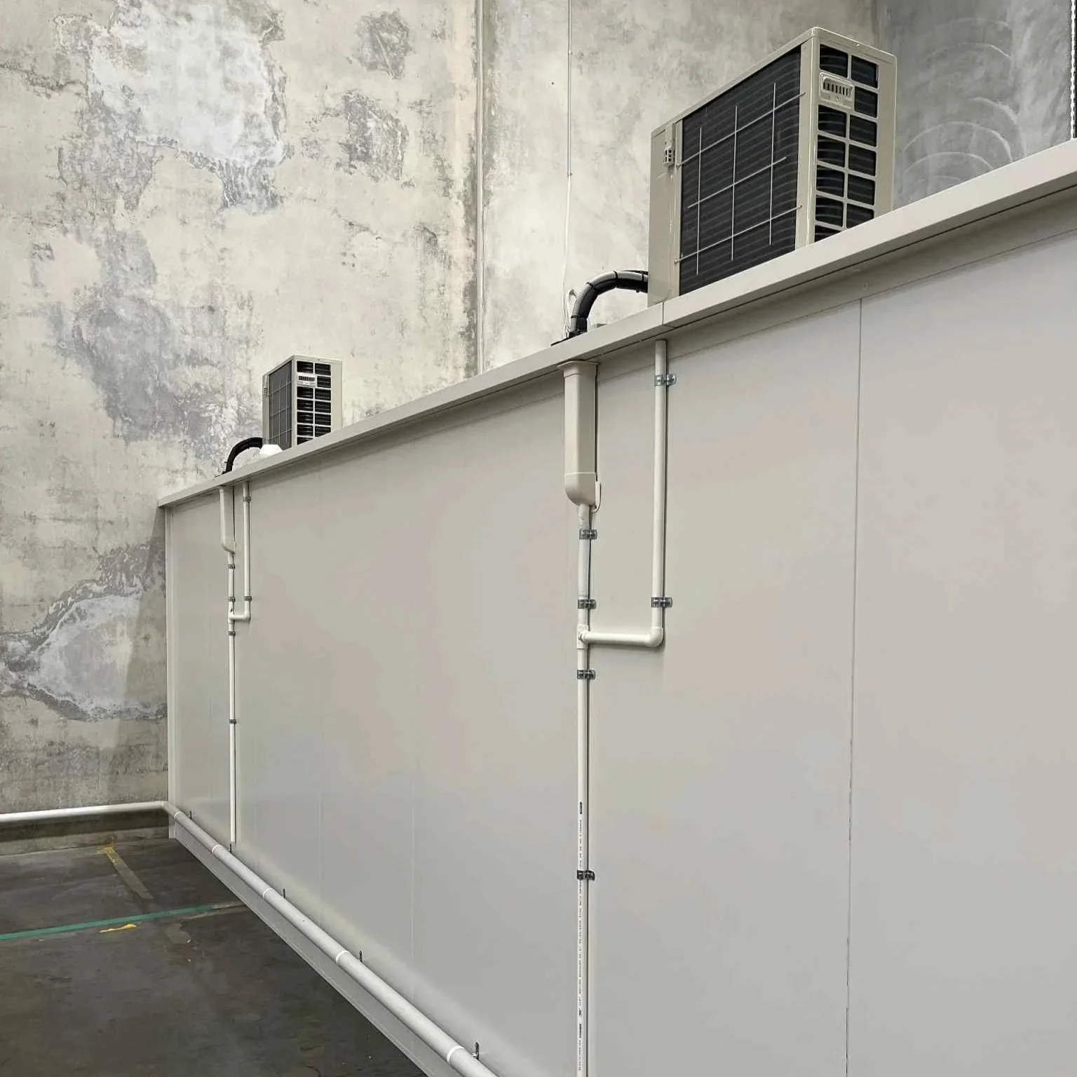 Two air conditioning units on top of a white structure with pipework against a concrete wall.