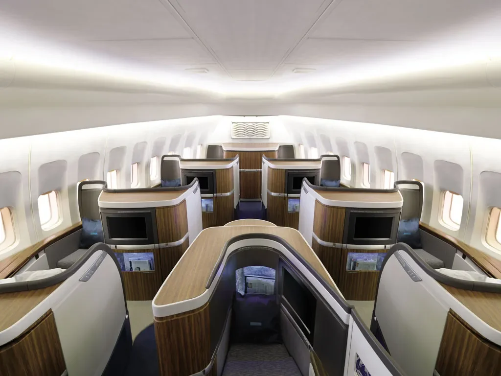 Interior of an airplane cabin with premium or business class seats arranged in a 2-2 configuration, featuring built-in screens, wood paneling, and large windows.