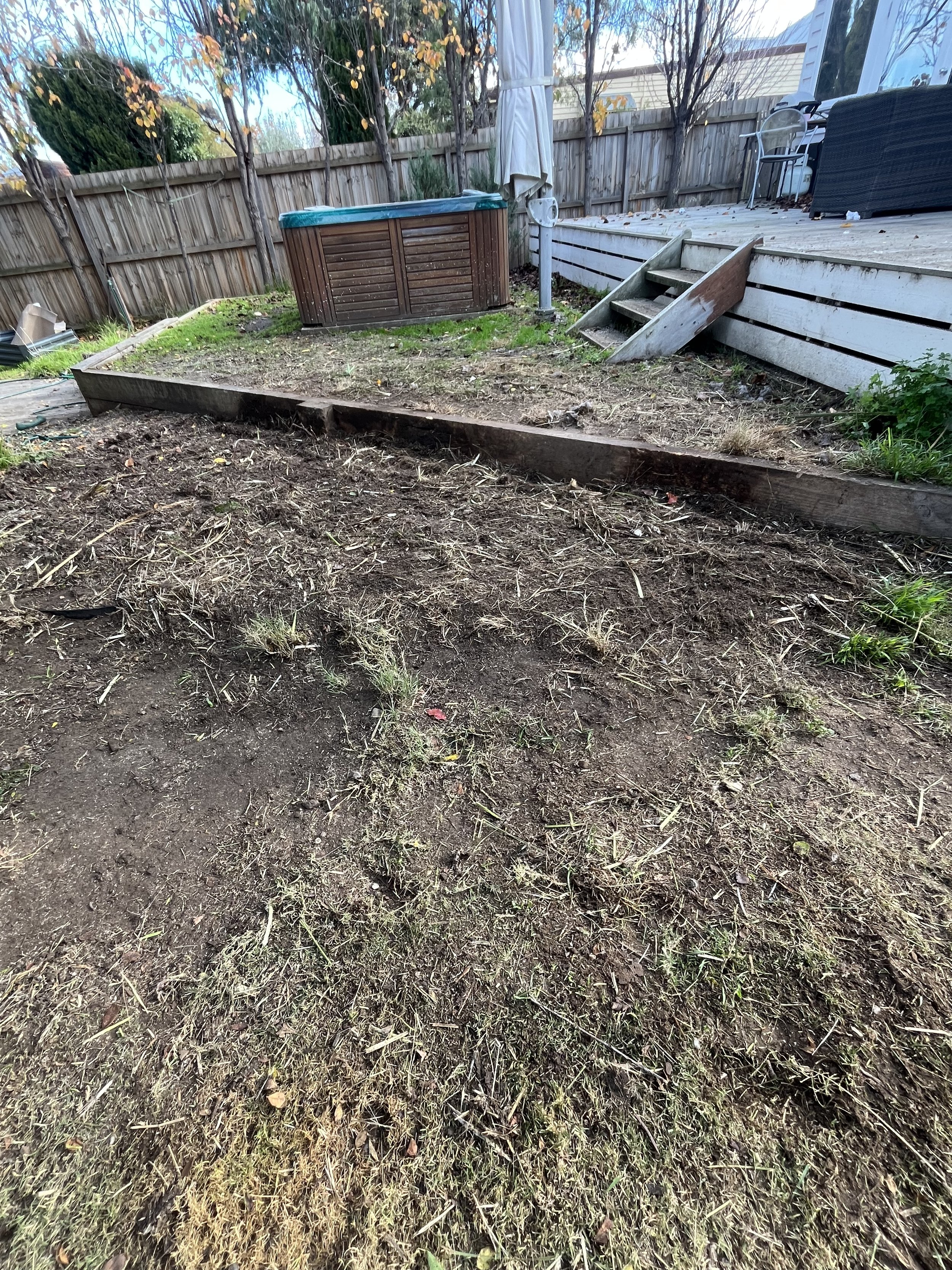 Backyard with a partially cleared garden bed, wooden retaining edges, deck stairs, hot tub, and outdoor furniture behind a wooden fence, during late fall.