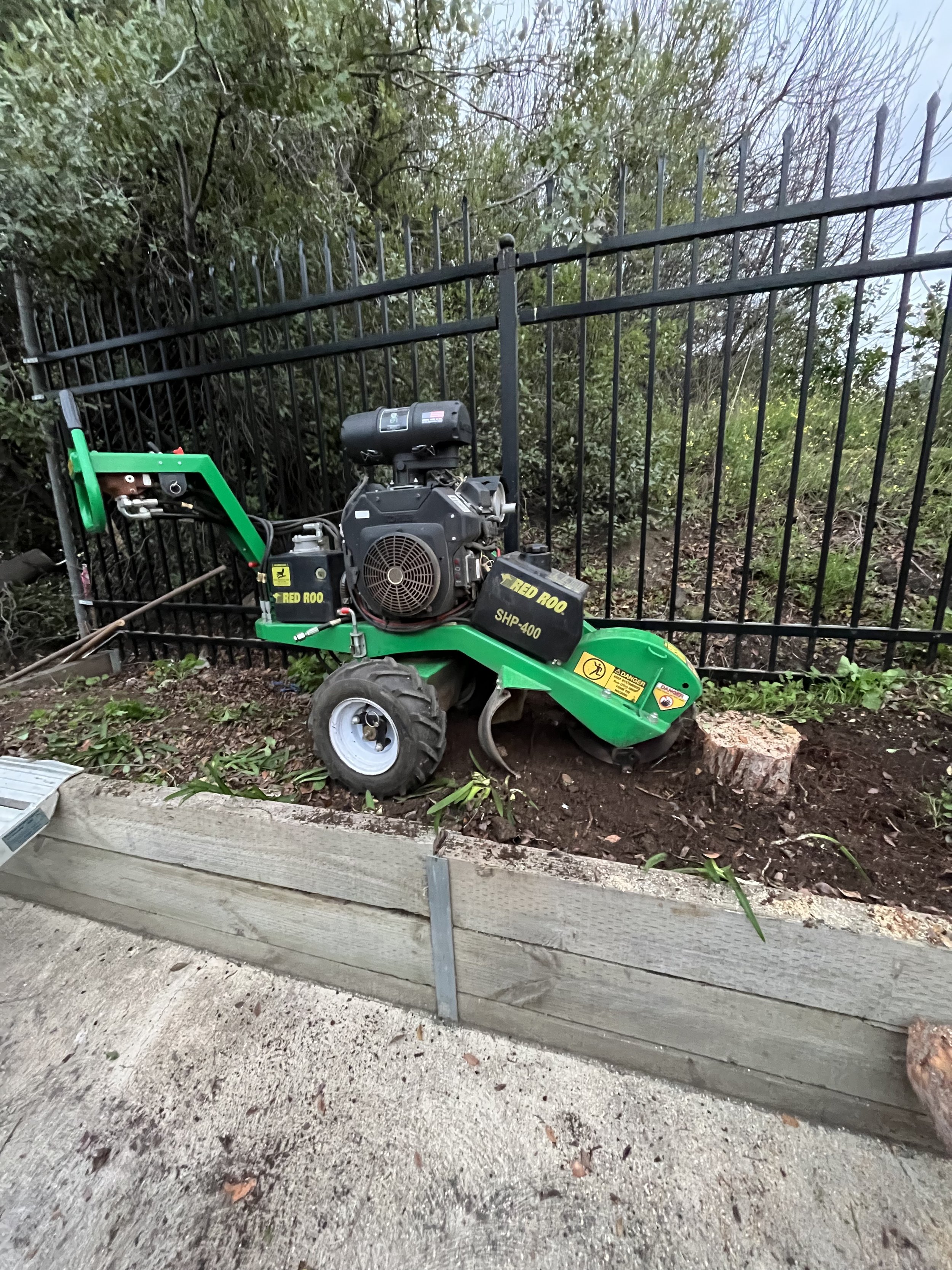 A tight access stump grinder machine positioned up on a retaining wall, next to a black metal fence, with a small side of soil, a tree stump in the foreground and next to a carpark