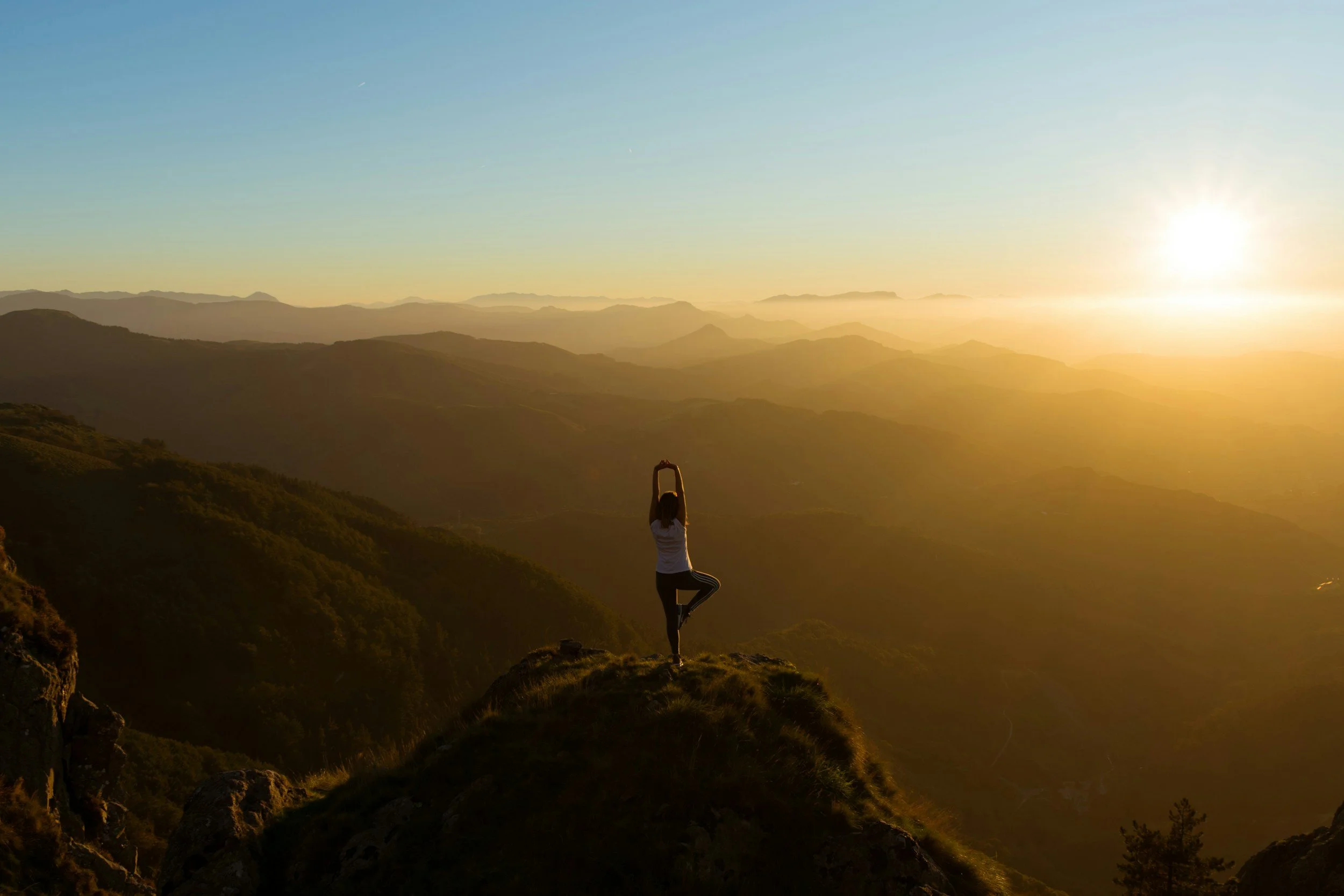 A person practicing yoga in a tree pose on a hilltop during sunset with a mountain landscape in the background.