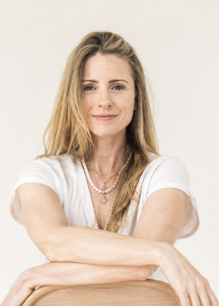 A woman with long blonde hair and blue eyes, wearing a white t-shirt and layered necklaces, sitting with her arms resting on a wooden surface, smiling softly at the camera.