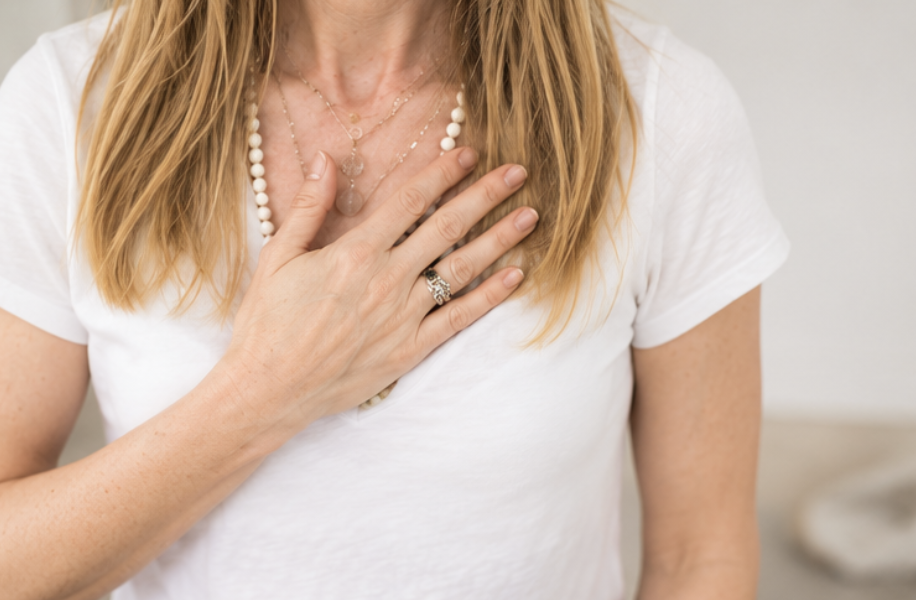 A woman with blonde hair, wearing a white shirt and layered necklaces, places her hand over her chest.