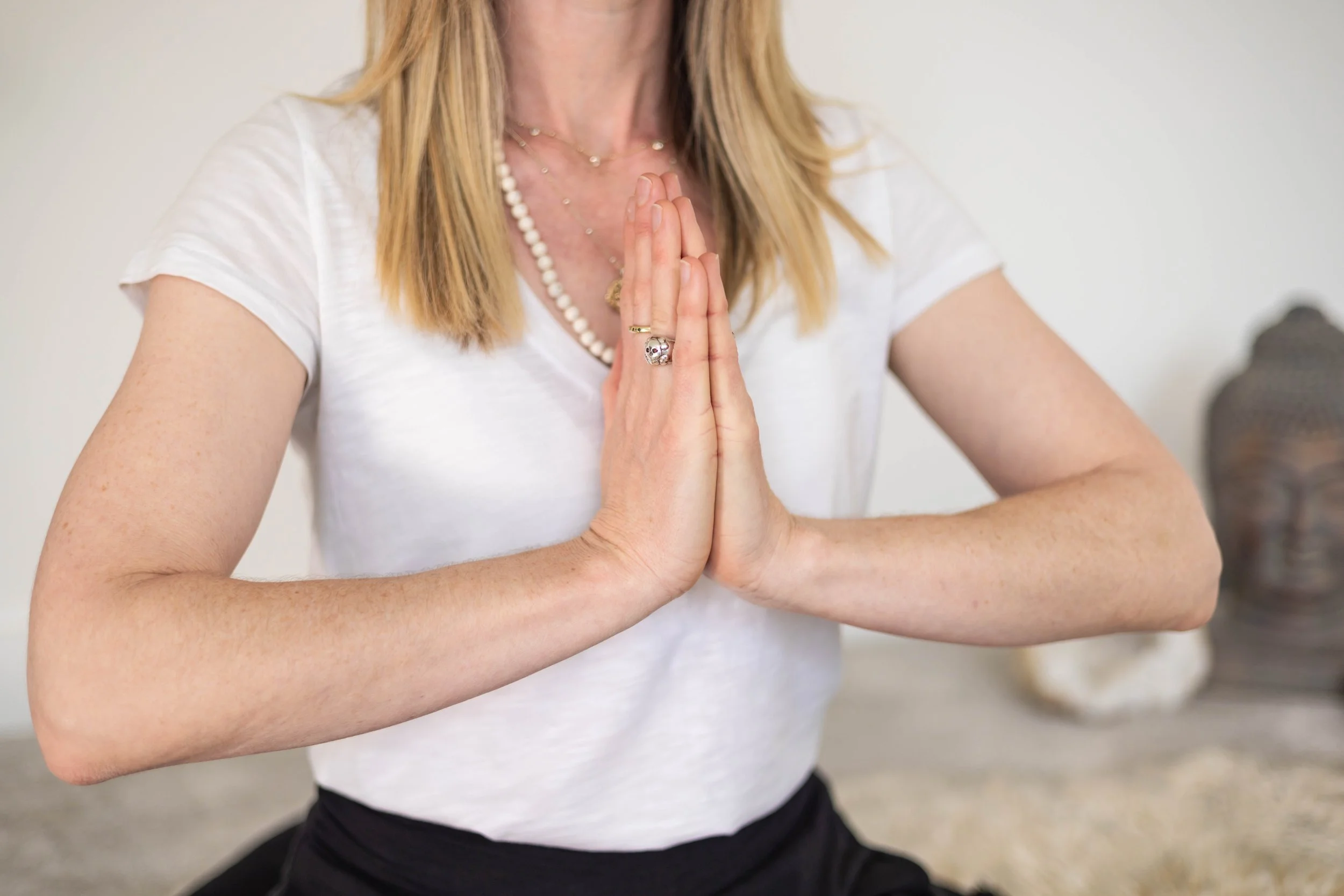 Woman practicing yoga, sitting cross-legged in a meditative pose with hands pressed together in front of her chest, wearing a white t-shirt and jewelry.