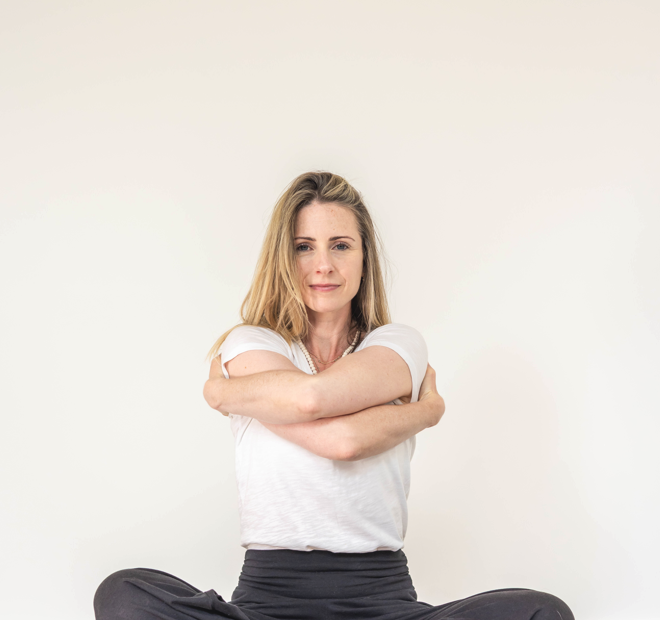 A woman sitting cross-legged on the floor with her arms wrapped around herself against a plain white background.