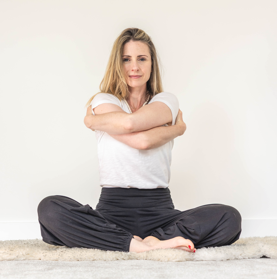 A woman sitting cross-legged on a carpet, hugging herself with her arms crossed, against a plain white wall background.