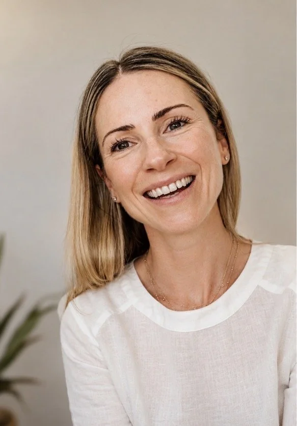 A woman with long blonde hair smiling at the camera, wearing a white top and small earrings.
