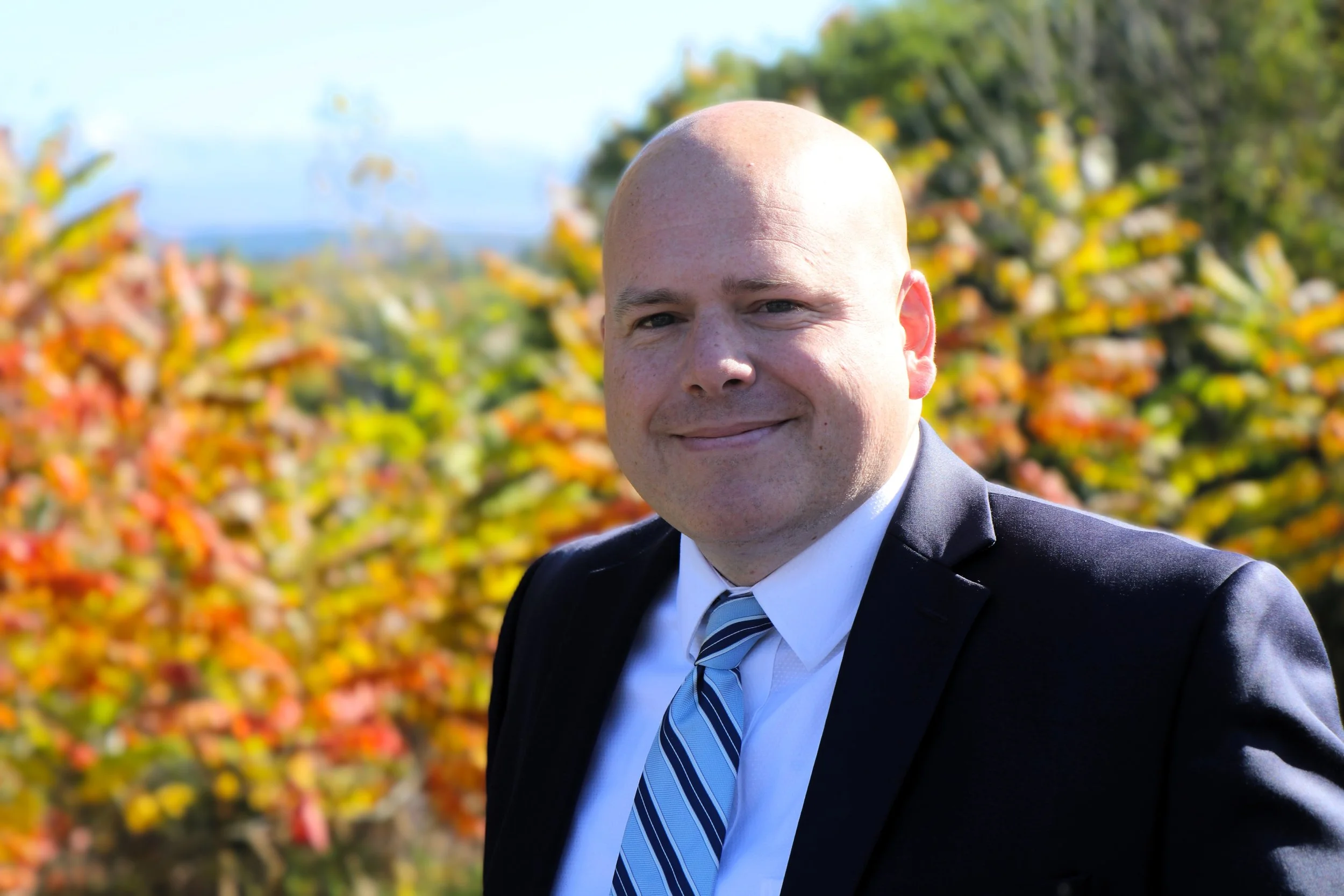 A smiling man with a bald head wearing a black suit, white shirt, and striped tie who looks like a lawyer outdoors during autumn in Vermont with colorful fall foliage in the background.