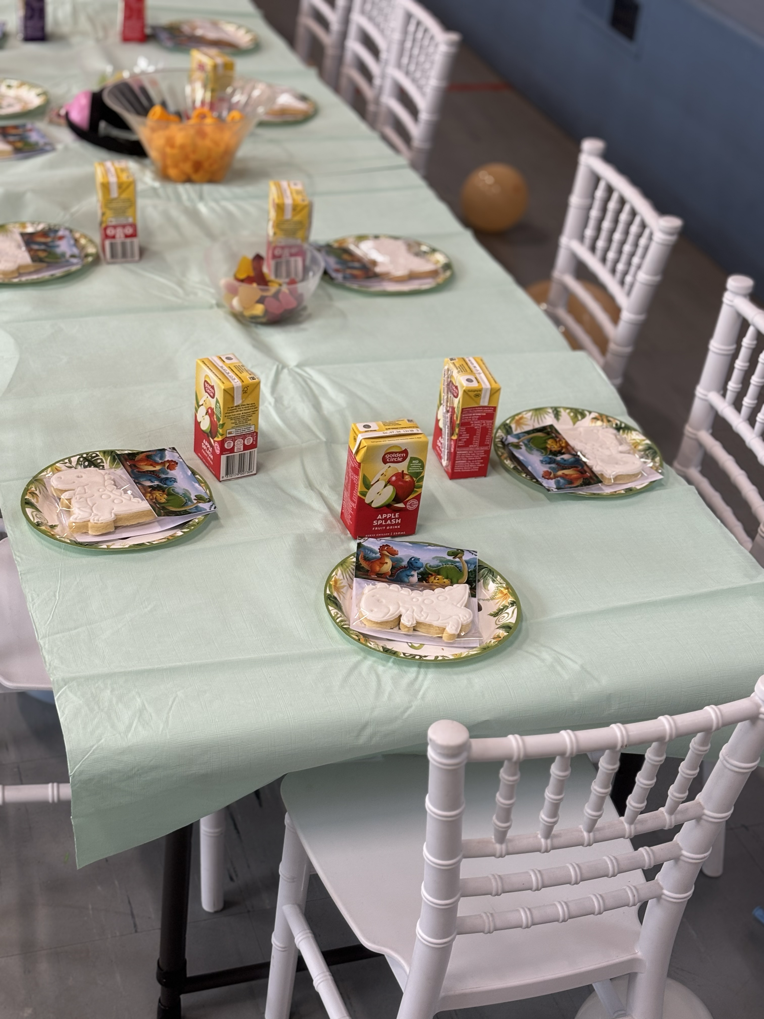Party table set with pastel green tablecloth, paper plates with a colorful cartoon design, White decorated cookies on plates, cartons of apple juice, a bowl of orange candies, and scattered paper napkins.