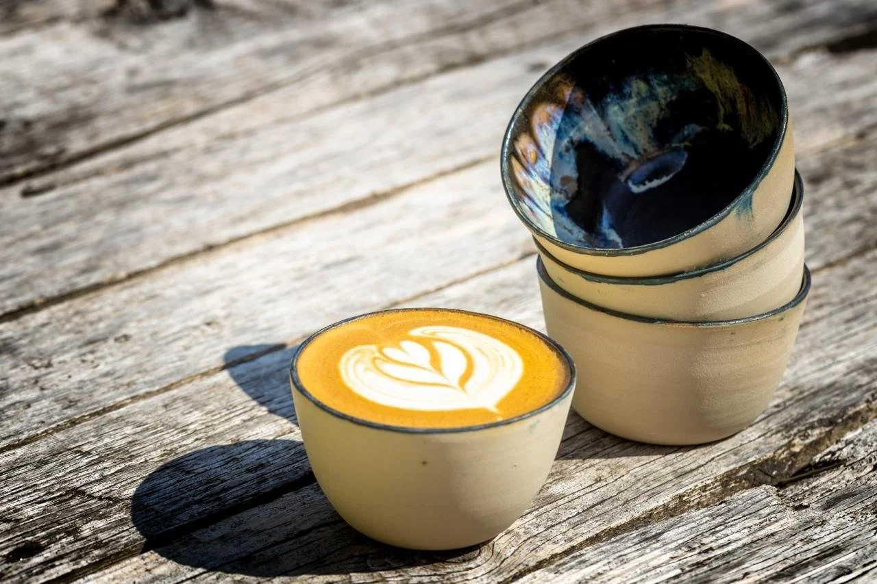 Three ceramic cups stacked and one cup with a latte art heart design on top, all placed on a weathered wooden surface outside.