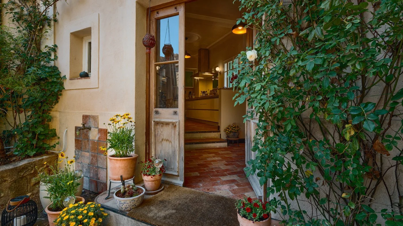 Open door to a cozy yellow kitchen with green plants and red flowers outside