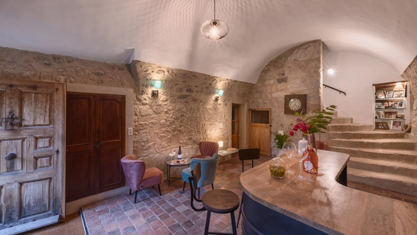 Interior of a cozy living room with stone walls, stairs, a wooden counter with flowers, bottles, and glasses, and a small seating area with colorful chairs.