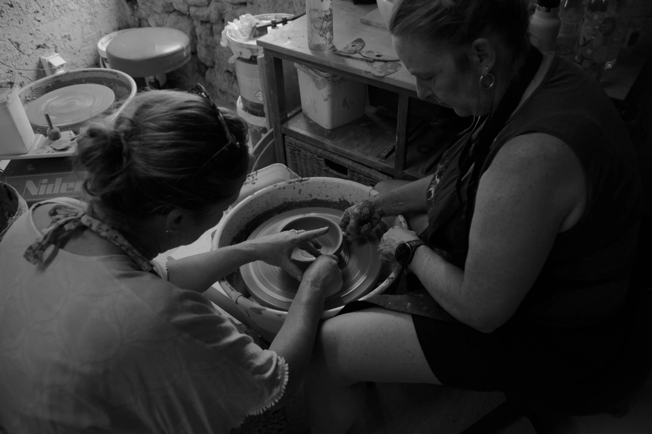 Two women working at a pottery wheel, shaping a ceramic piece together in a ceramics studio.