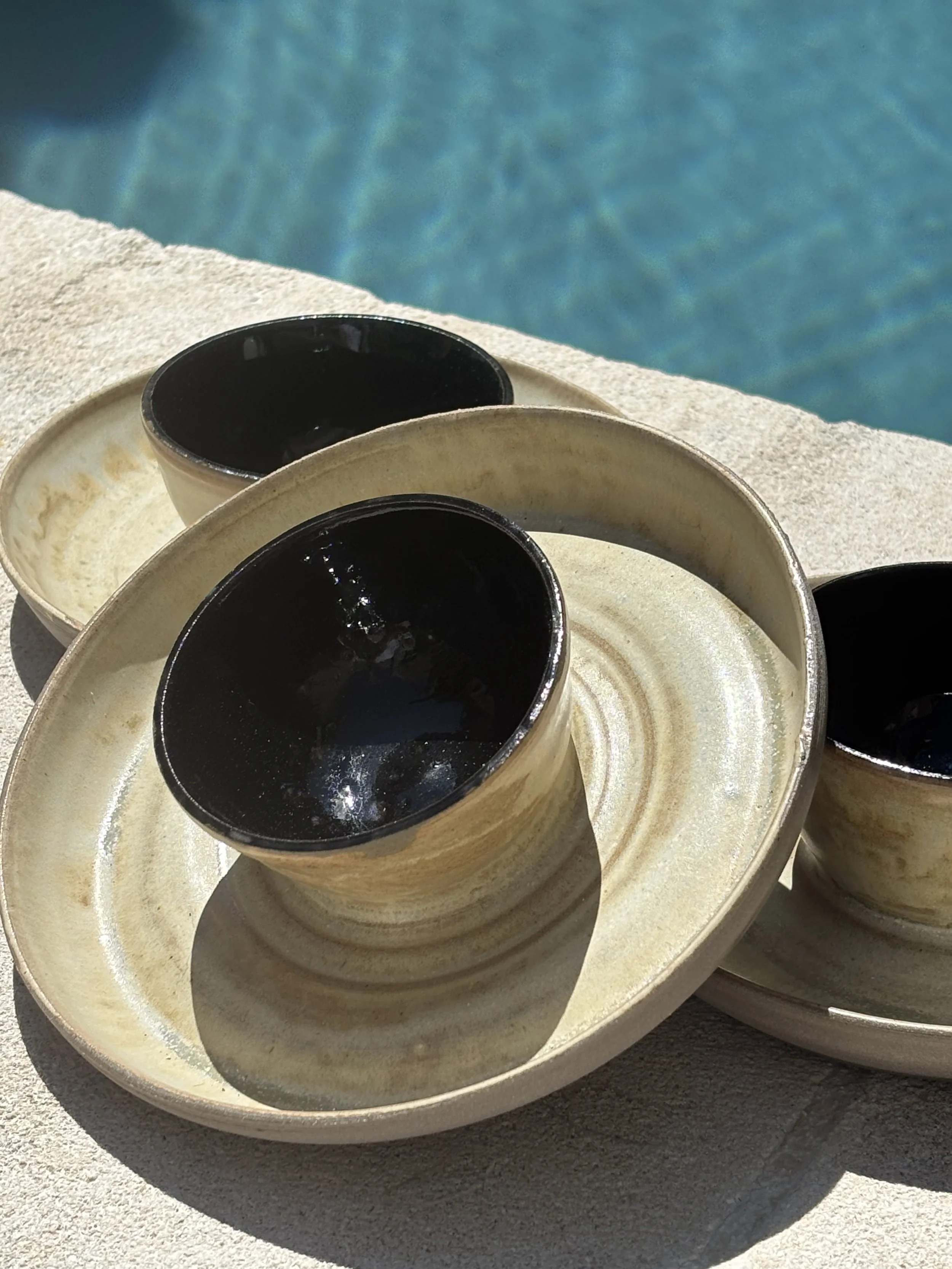 Small ceramic cups filled with soy sauce on beige trays, set on a poolside surface with a swimming pool in the background.
