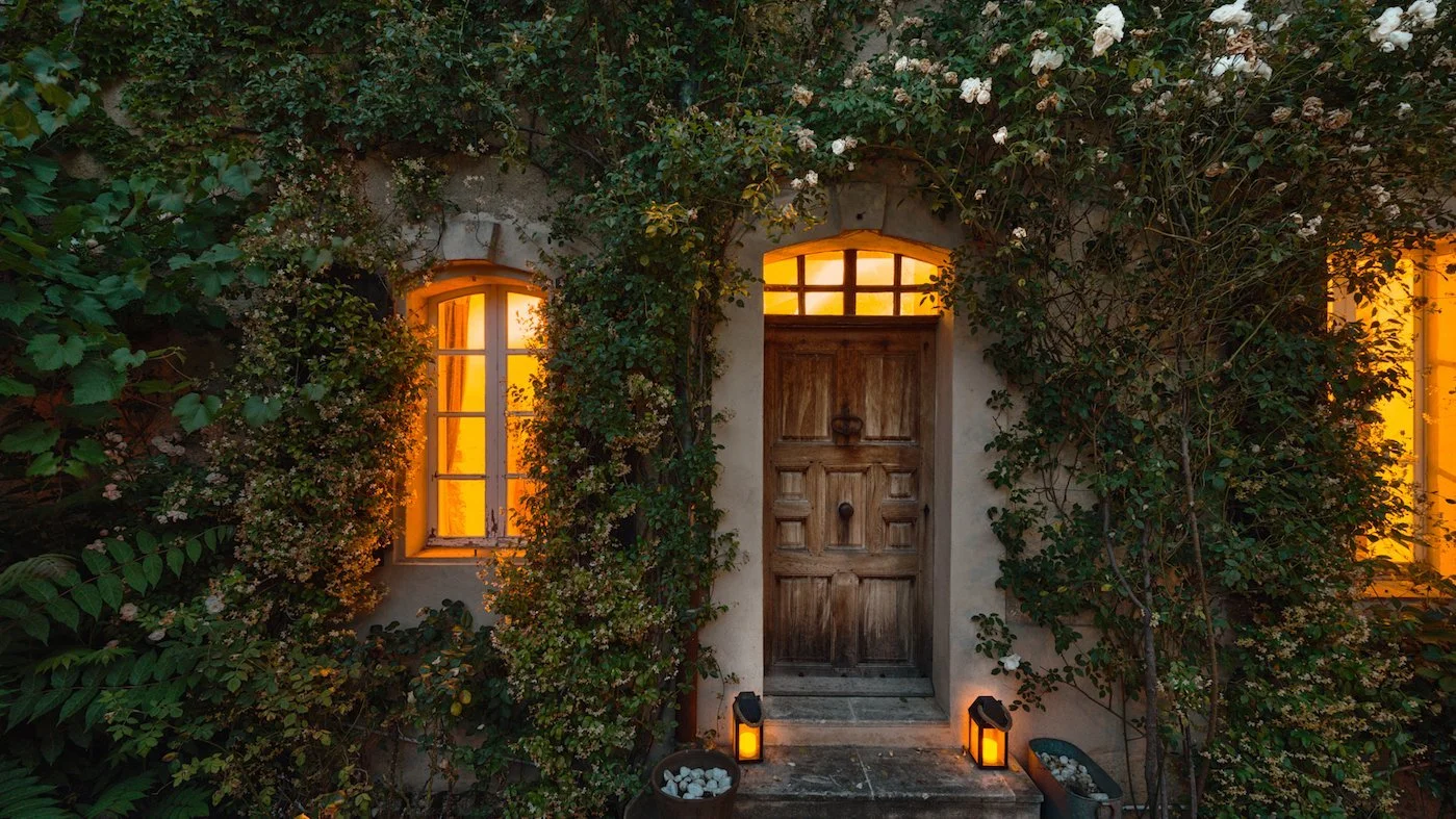 A rustic wooden front door with glow-in-the-dark lanterns on a stone step, surrounded by green vines and white flowers, with warm light coming from windows.