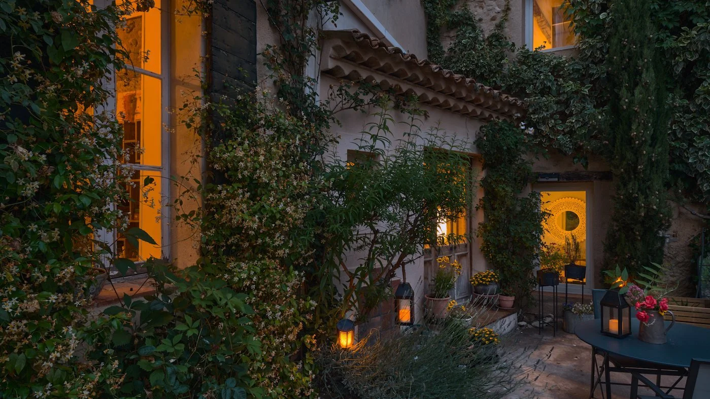 A cozy outdoor patio decorated with potted plants, lanterns, and string lights, with a view into a warm-lit house interior.