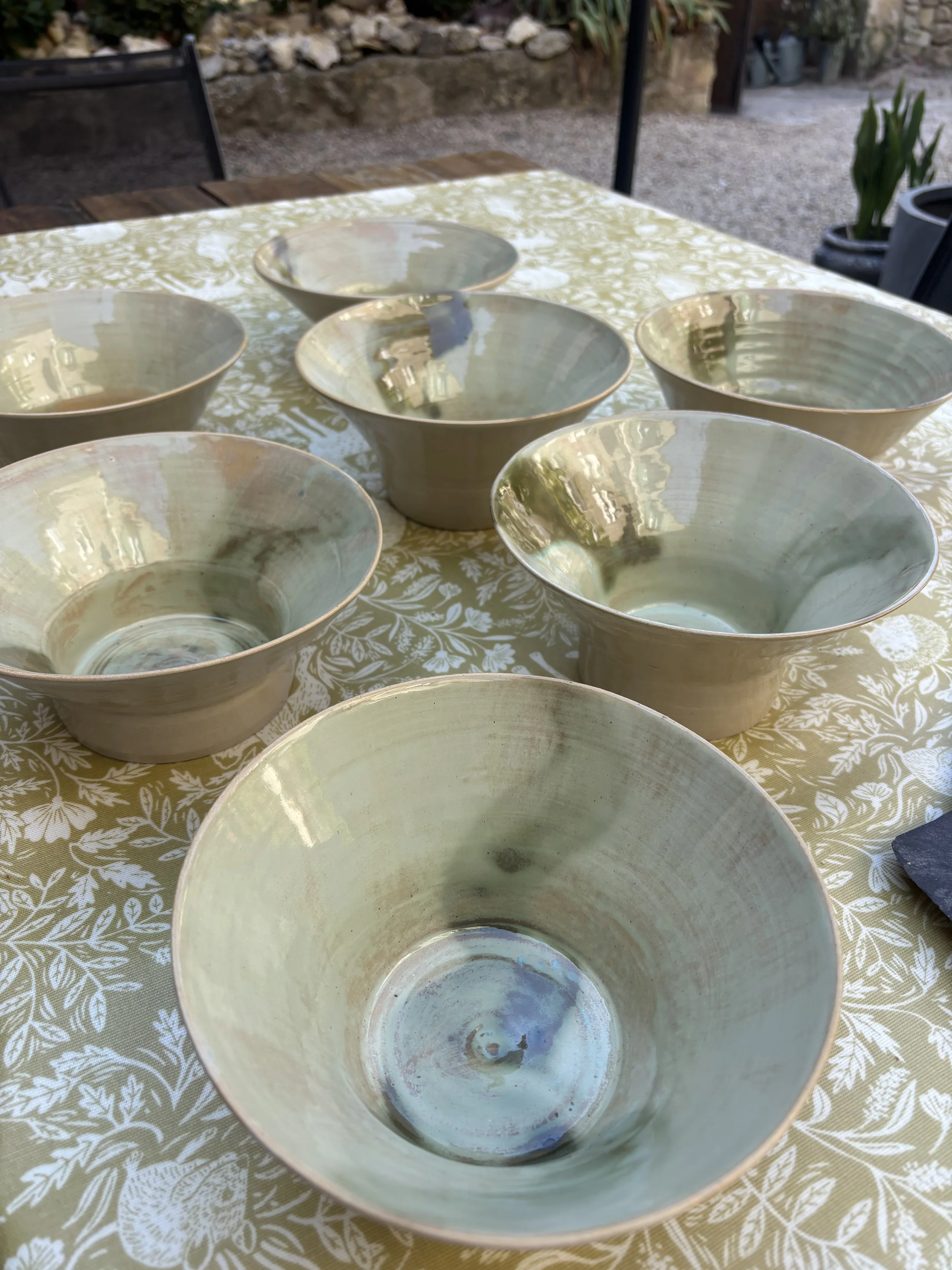 Several light-colored ceramic bowls placed on a patterned yellow tablecloth outdoors, with a stone wall and plants in the background.