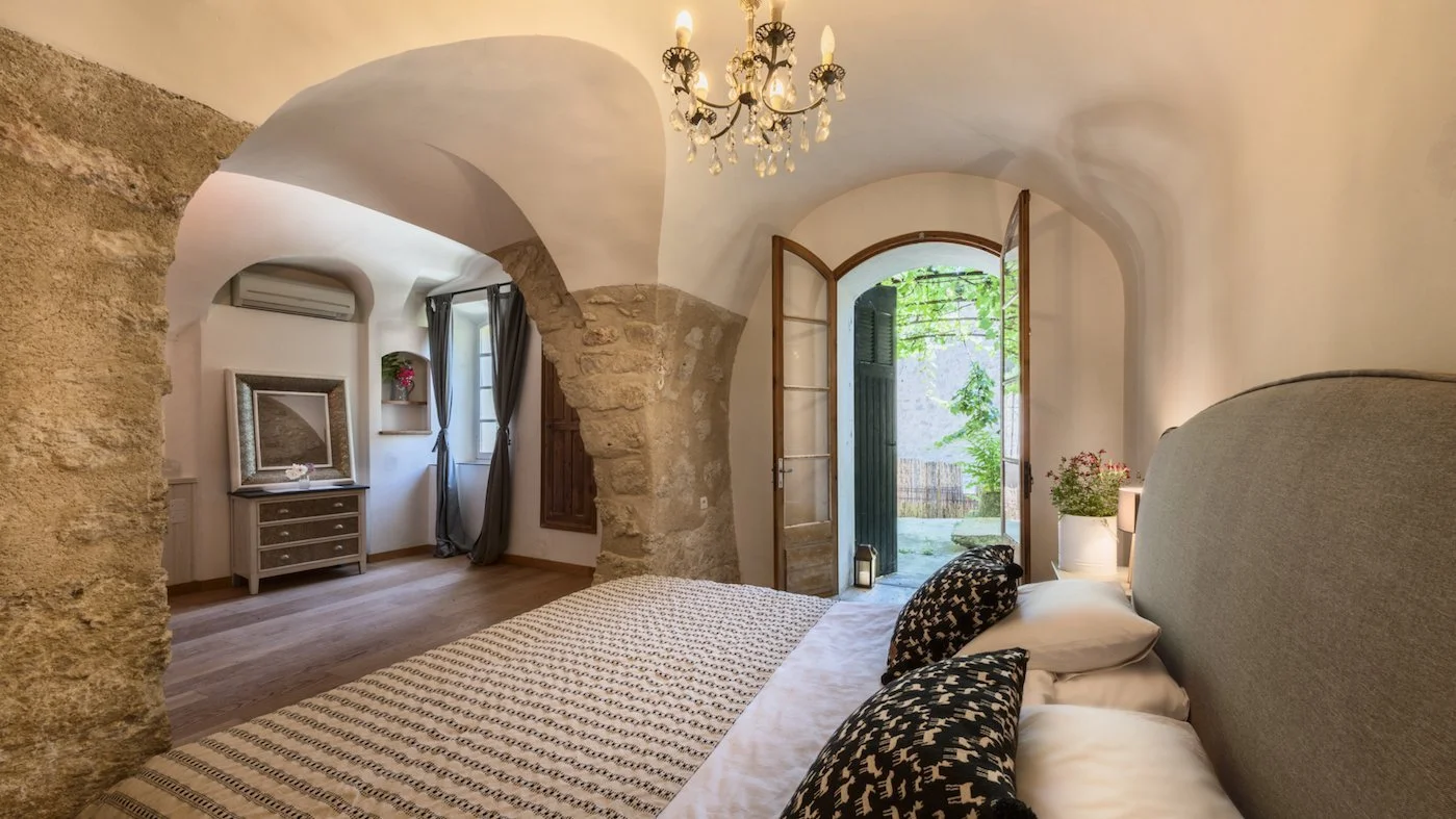 Cozy bedroom with stone and plaster walls, a bed with white and black patterned pillows, a chandelier, and an open door leading to a garden.