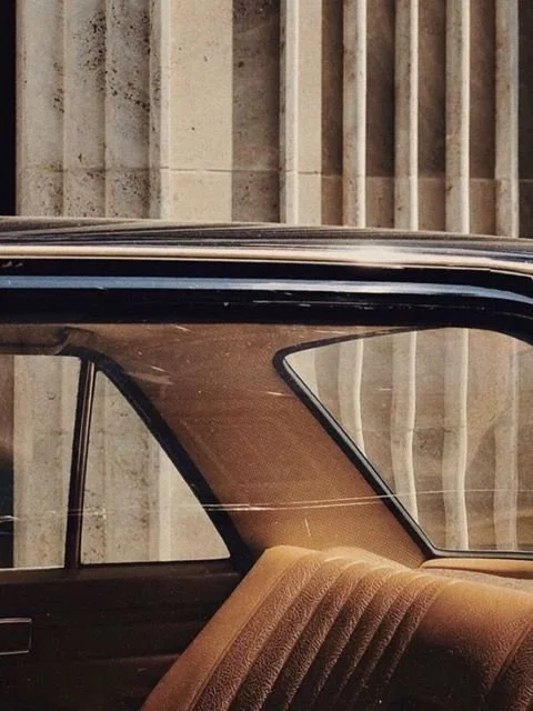 Close-up of the interior of a vintage car, showing part of the front seat and the side window with a concrete wall in the background.