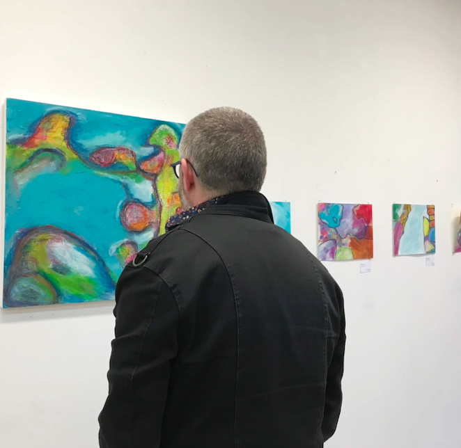 A man with short, gray hair and glasses wearing a black coat viewing Michelle Saleeba's abstract colourful paintings in an art gallery.