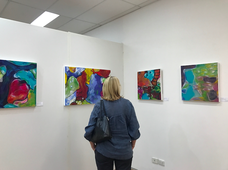 A woman with blonde hair in a blue jacket carrying a black handbag observing colourful abstract paintings by Michelle Saleeba on white gallery walls.