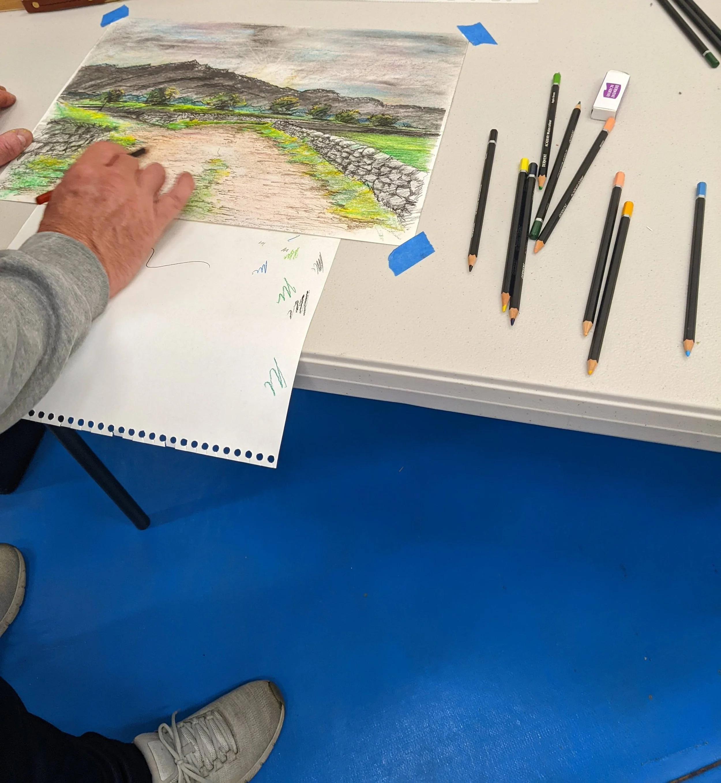 Mans hands drawing a landscape in coloured pencils participating in a wellbeing art group