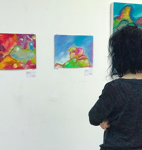 A woman with curly black hair and a dark sweater stands with her arms crossed, contemplating colourful abstract paintings by Michelle Saleeba on a white gallery wall.