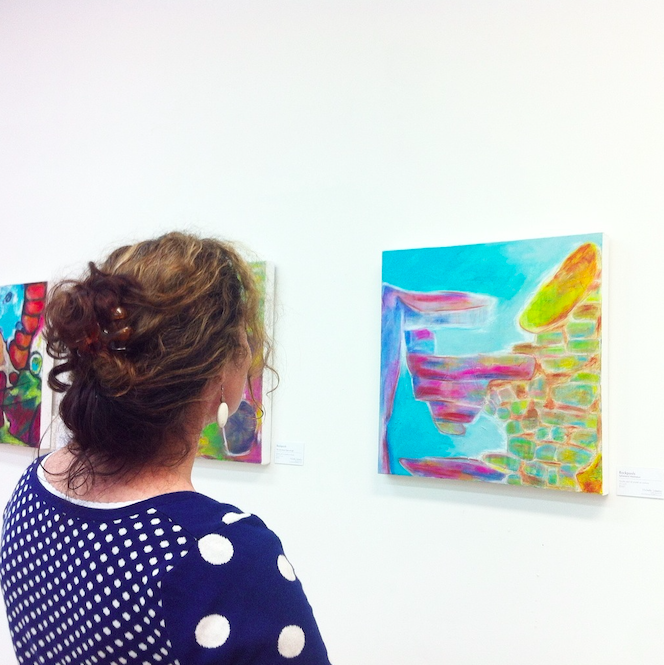 A woman with curly hair wearing a blue sweater with white polka dots is looking at colourful abstract paintings in an exhibition of artwork by artist Michelle Saleeba