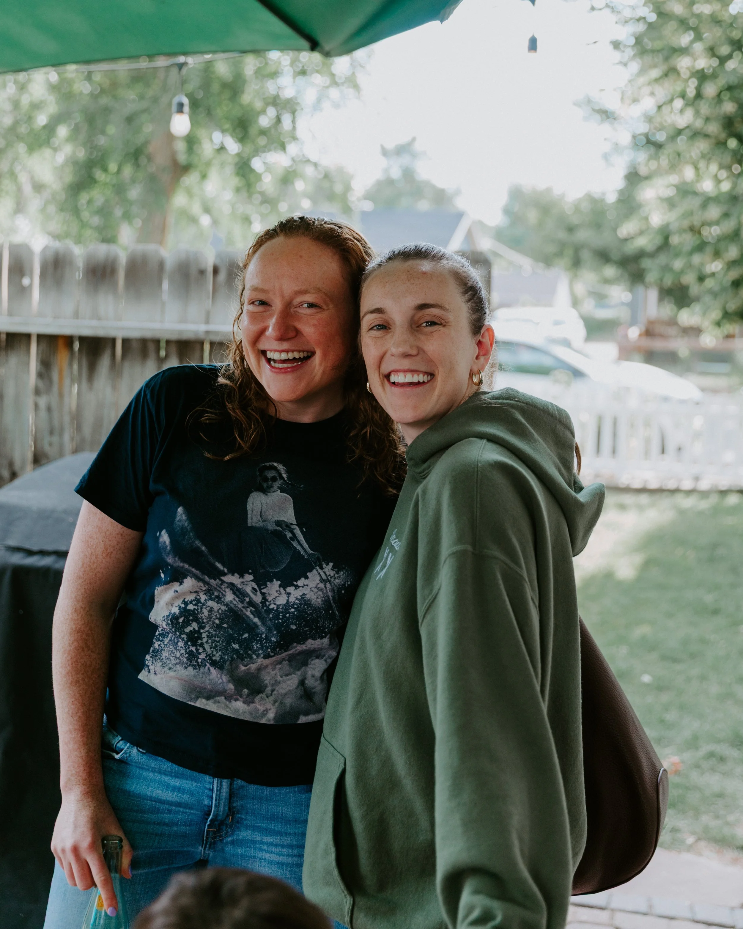 Two young women smiling and posing outdoors on a sunny day, one with curly red hair wearing a black t-shirt, and the other with brown hair in a green hoodie.