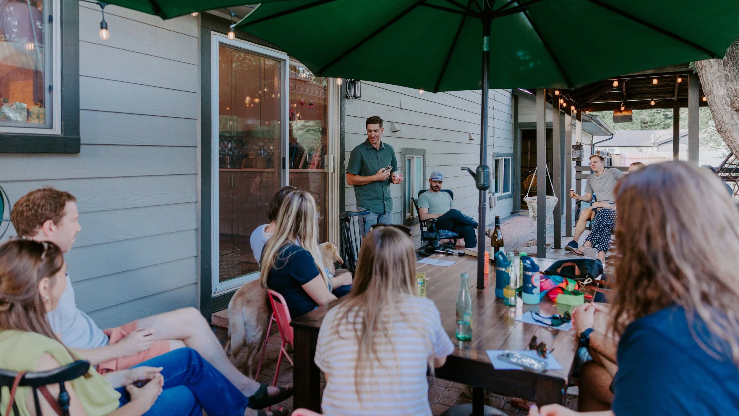 Outdoor gathering on a porch with friends, string lights, a large green umbrella, and a speaker, with a man standing and speaking to seated guests.