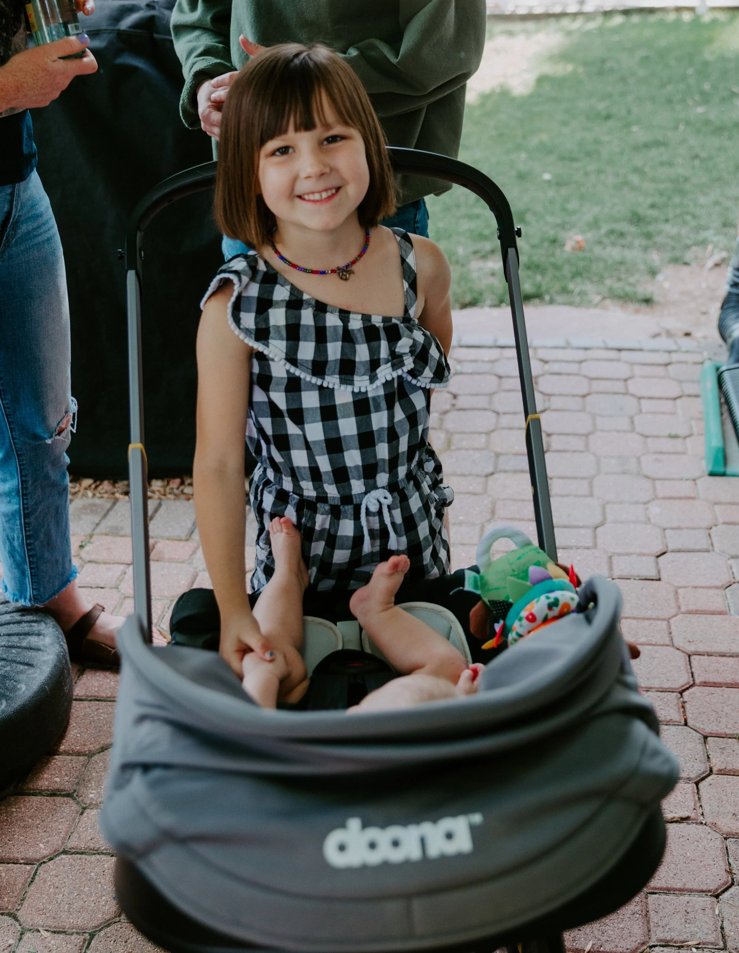 A young girl with shoulder-length brown hair, wearing a black and white checkered dress, is smiling while standing behind a gray Doona stroller with a baby inside. The baby is lying on their back, holding a toy, and has their feet up in the air. Ther