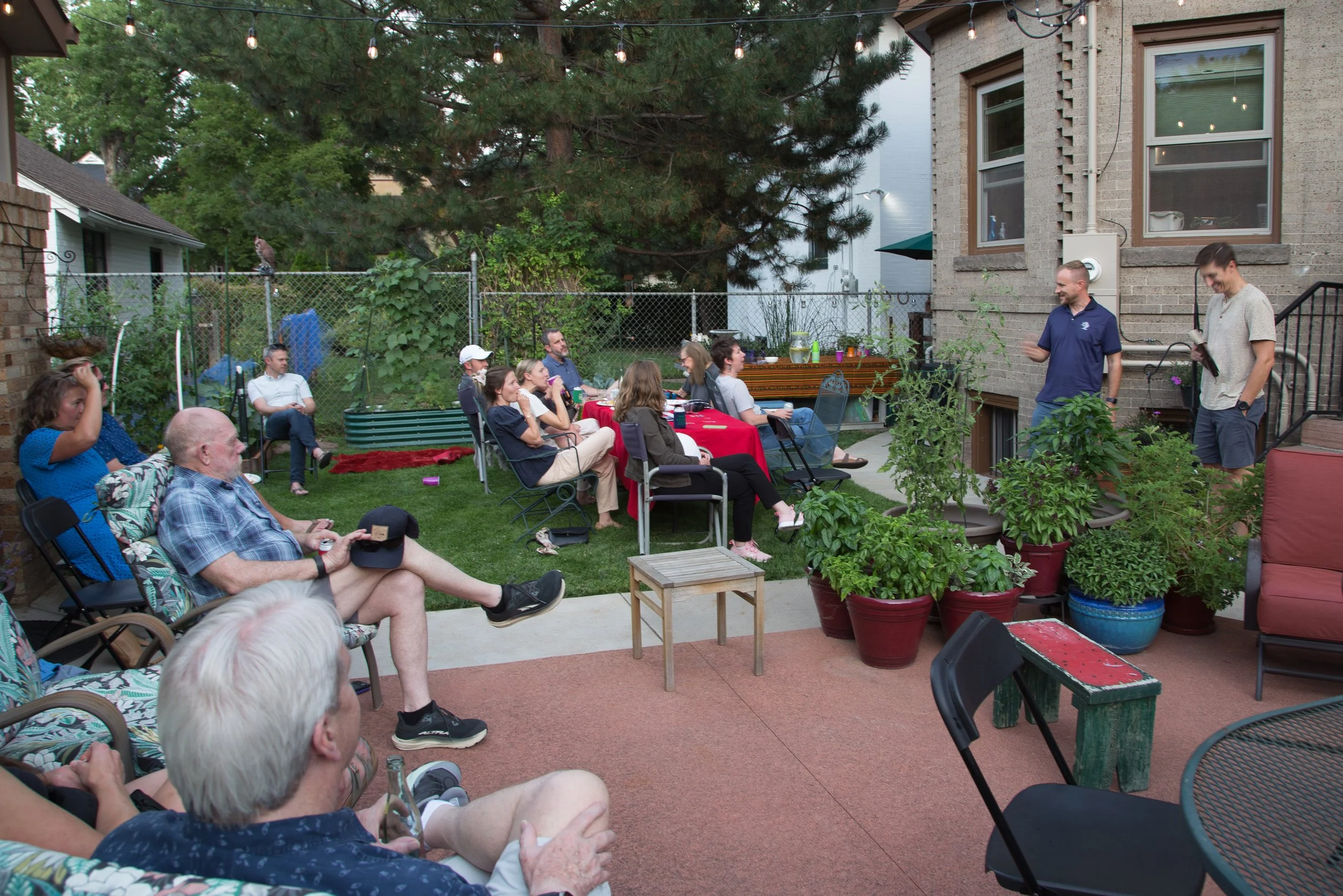 People gathered in a backyard for a social gathering or party during the daytime, sitting on chairs and benches, with two men standing and speaking to the group near a house, and plants and string lights decorating the area.