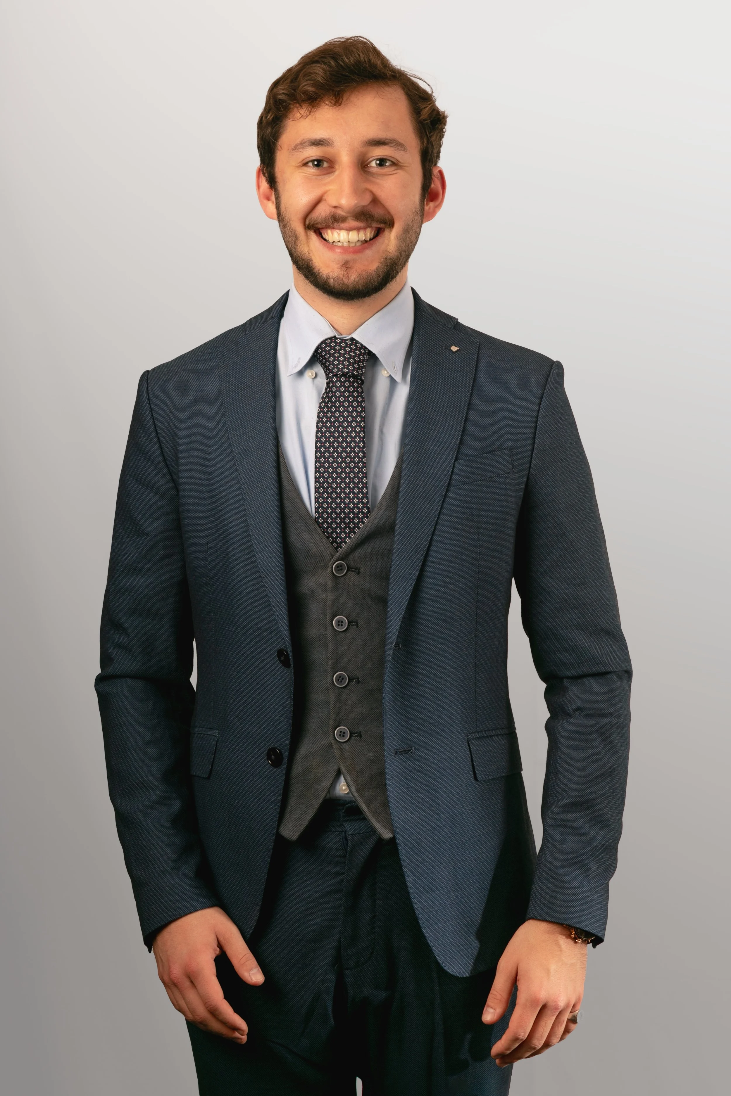 A young man with brown hair and a beard, smiling, in a navy blue suit, white shirt, and tie, standing against a plain light-colored background.
