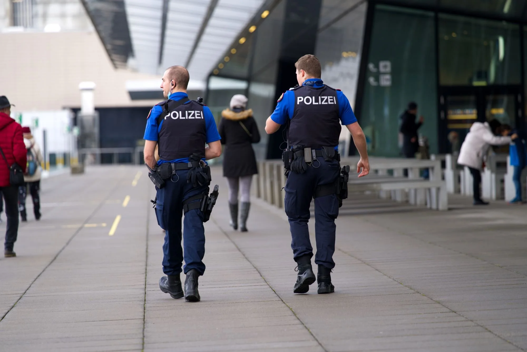 Two police officers in uniforms walking outside a modern building, with other people around.